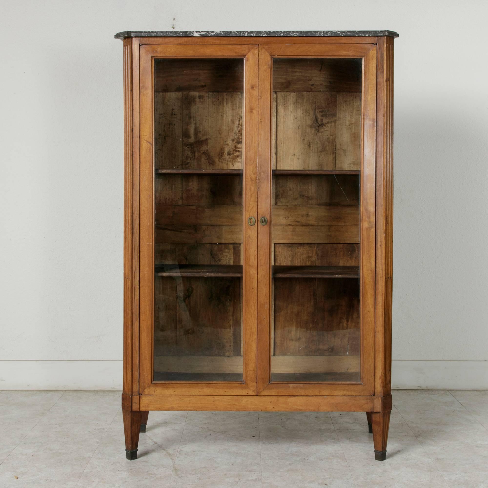 This Louis XVI period walnut bibliotheque or vitrine features fluted corners that lead to tapered feet finished in bronze sabots. Each side is constructed of a solid walnut panel made from a single board. A Saint Anne beveled marble top and locking