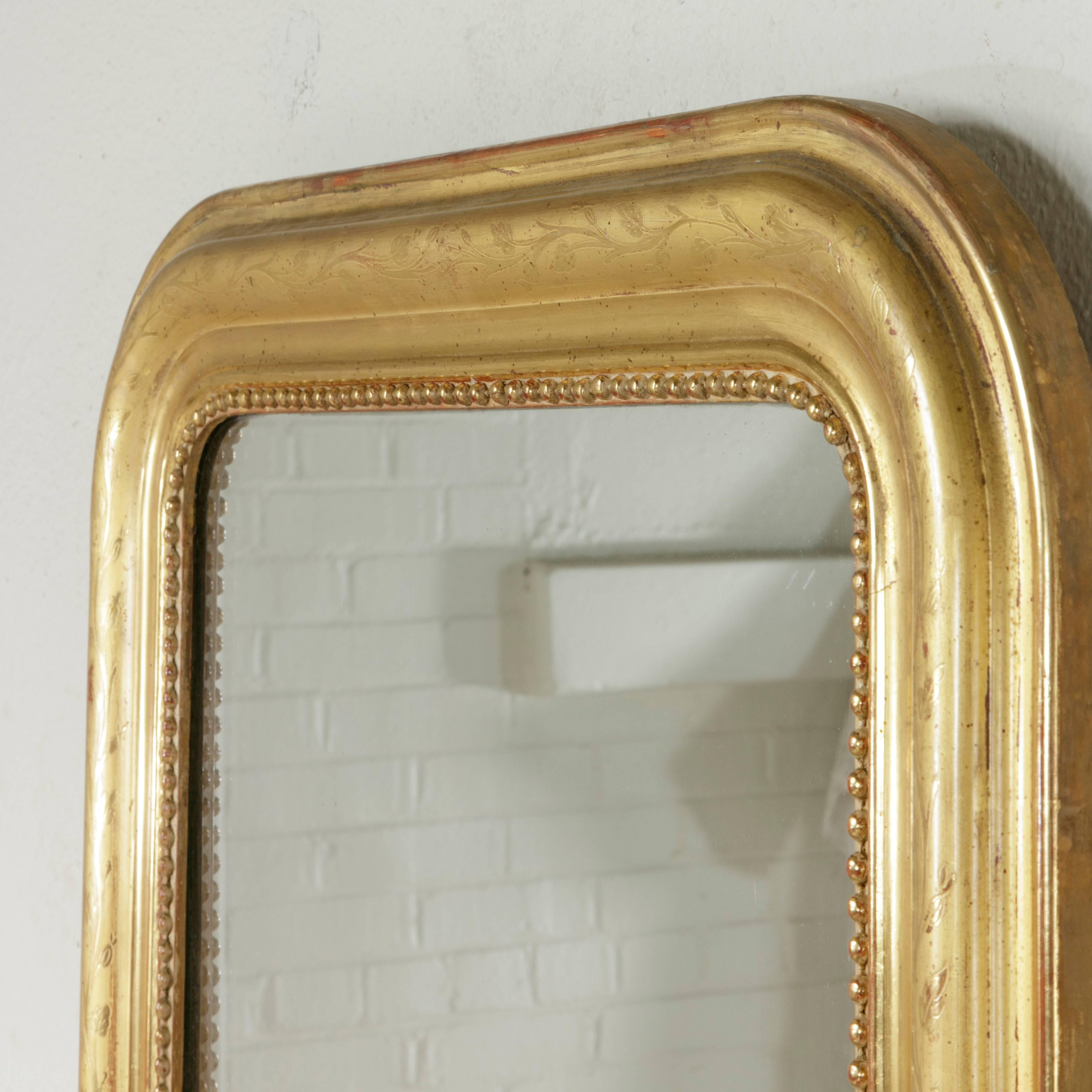 This elegant 19th century French Louis Philippe mirror features a delicately incised leaf and floral pattern on its gold leafed water gilt pillow frame. Its original iron oxide underbody gives it a rich glow. The original mercury glass mirror is