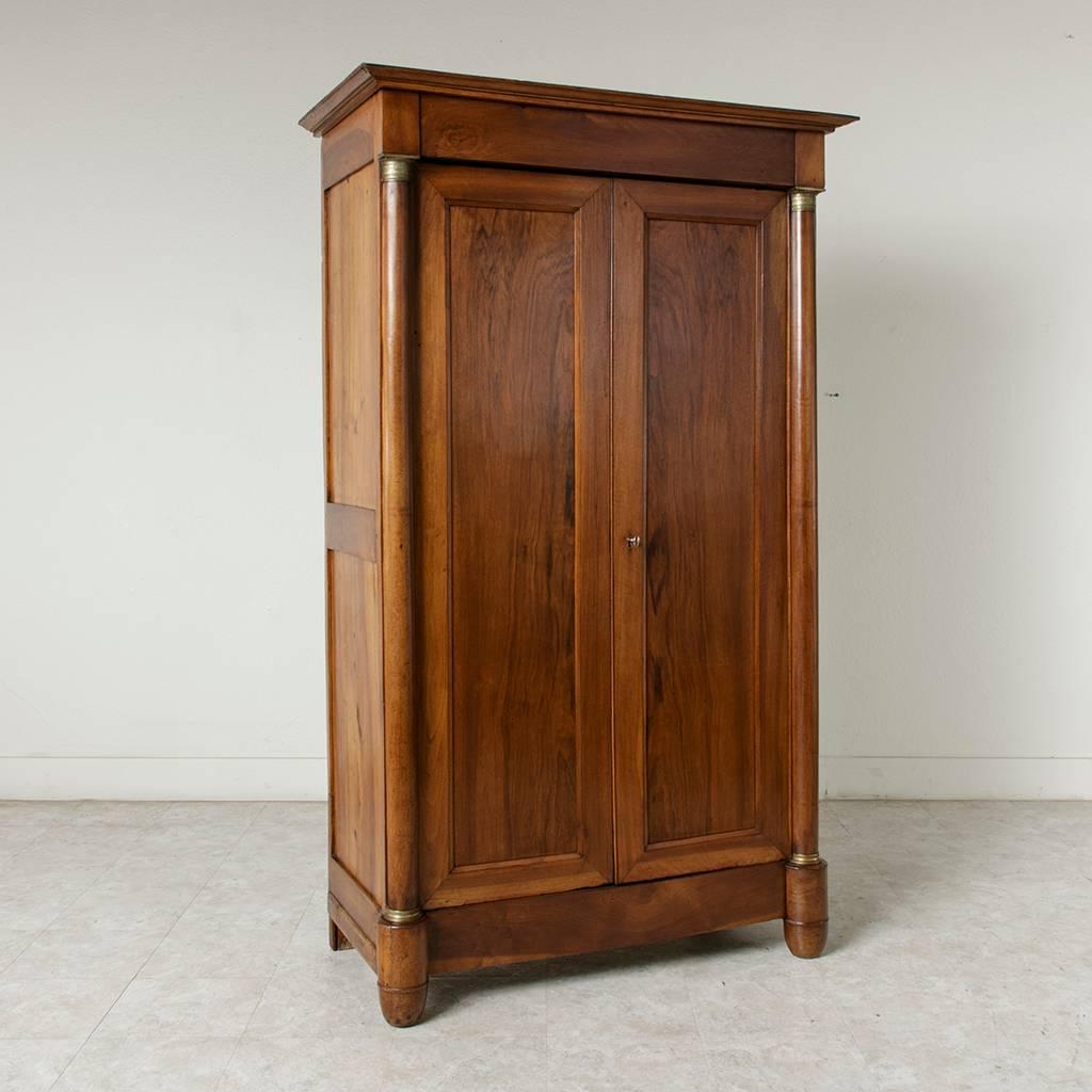 This small-scale walnut French Empire period armoire features double doors flanked by half columns that are fitted in bronze at the bases and capitals. Its sides are constructed of solid walnut. With three interior shelves and two parallel brass