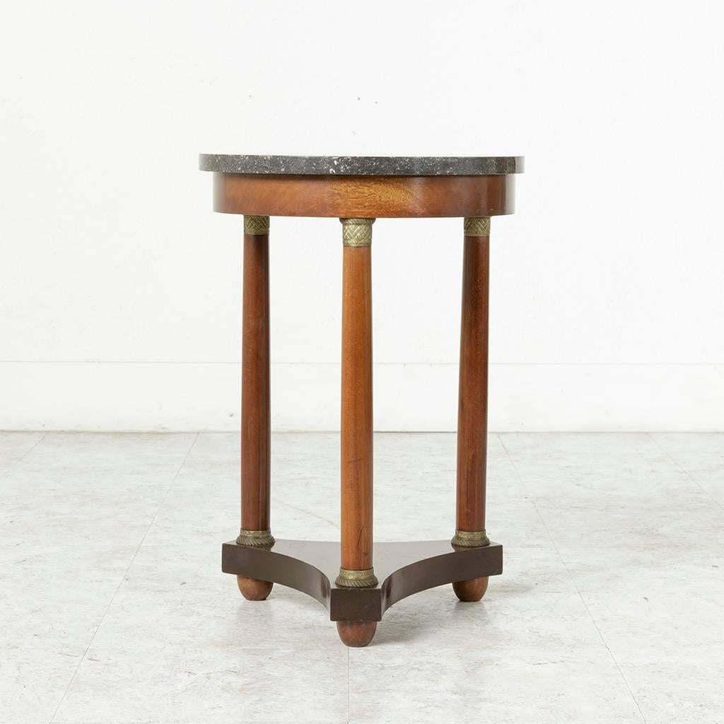 This French Empire style side table from the early 20th century features a mahogany base supported by three columns finished with bronze banding at their bases and capitals. Its lower level in the form of a pseudo-triangle with curved sides rests on
