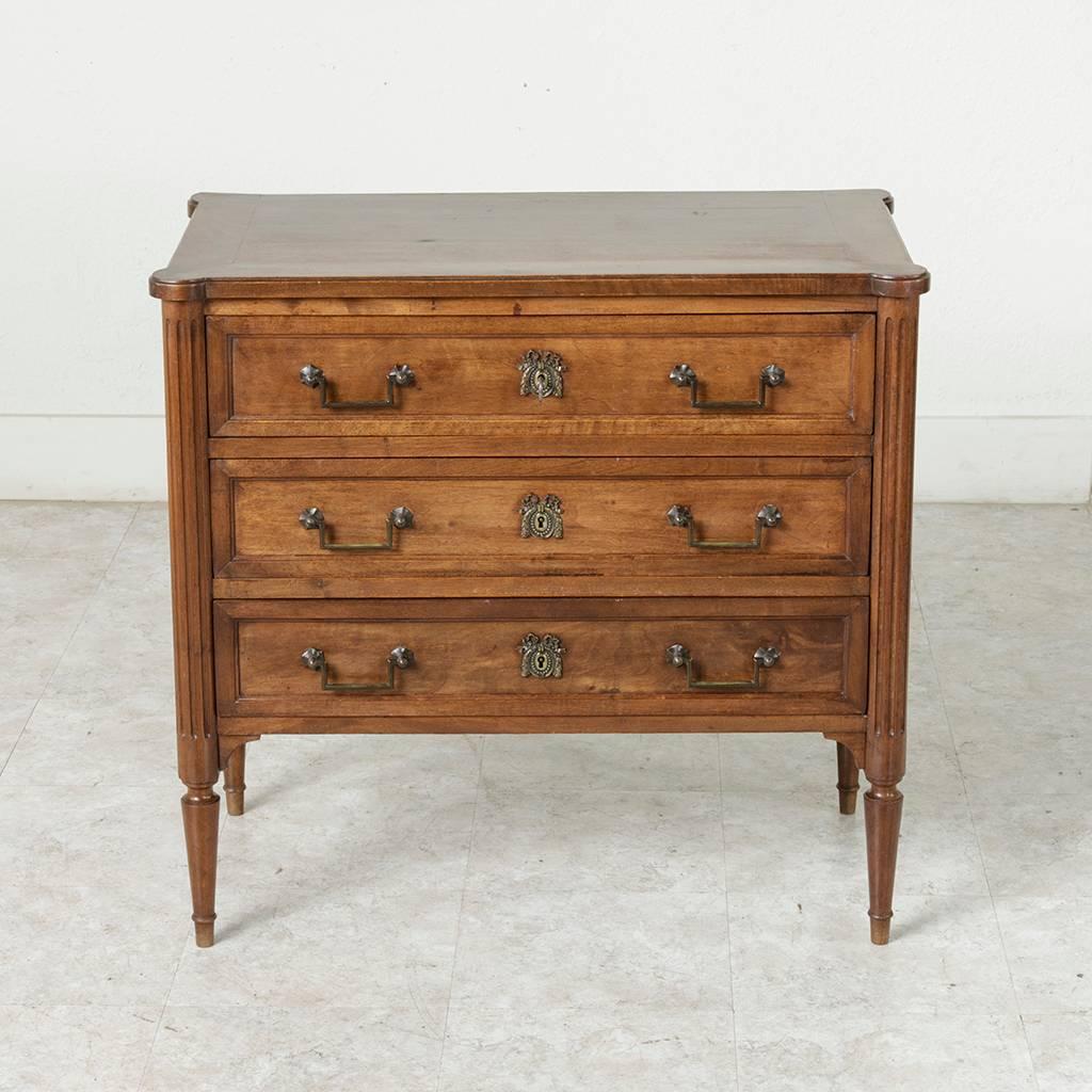A charming small-scale chest in the Louis XVI style, this early 20th century petite walnut commode features a bevelled top with rounded corners that rest on fluted legs leading to tapered feet. Three drawers of dovetail construction are detailed