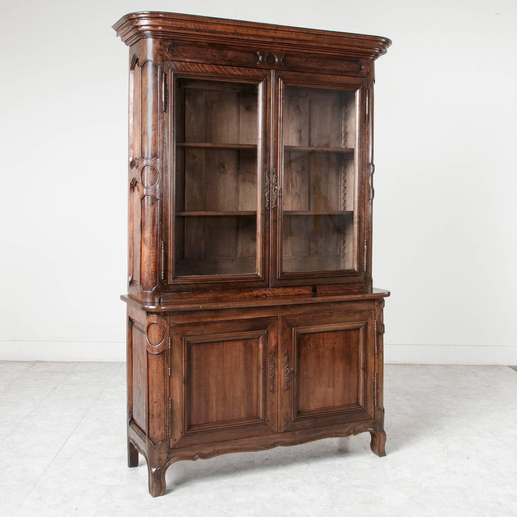 Unusual for its narrow 15-inch depth and exceptional deep relief on its hand-carved solid paneled sides, this 18th century hand pegged walnut bibliothèque, vitrine, or bookcase features handblown glass in the upper doors. Fitted with iron fiche