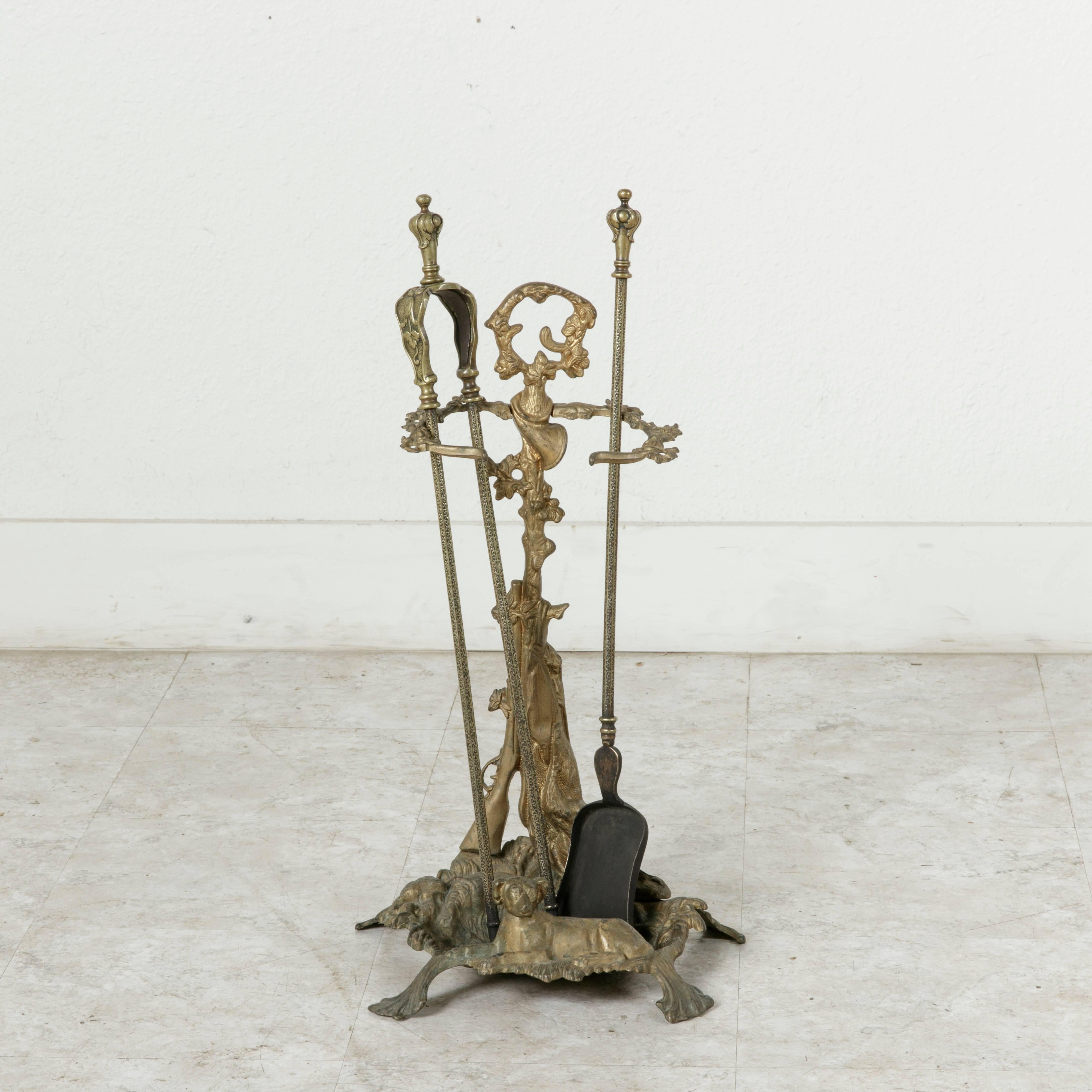 This set of fireplace tools includes its bronze stand with hunting motif, a shovel, and a pair of tongs. The stand features a hunting dog with a head that turns, lying down at the front of the base. Behind him a hunting rifle leans upon a tree