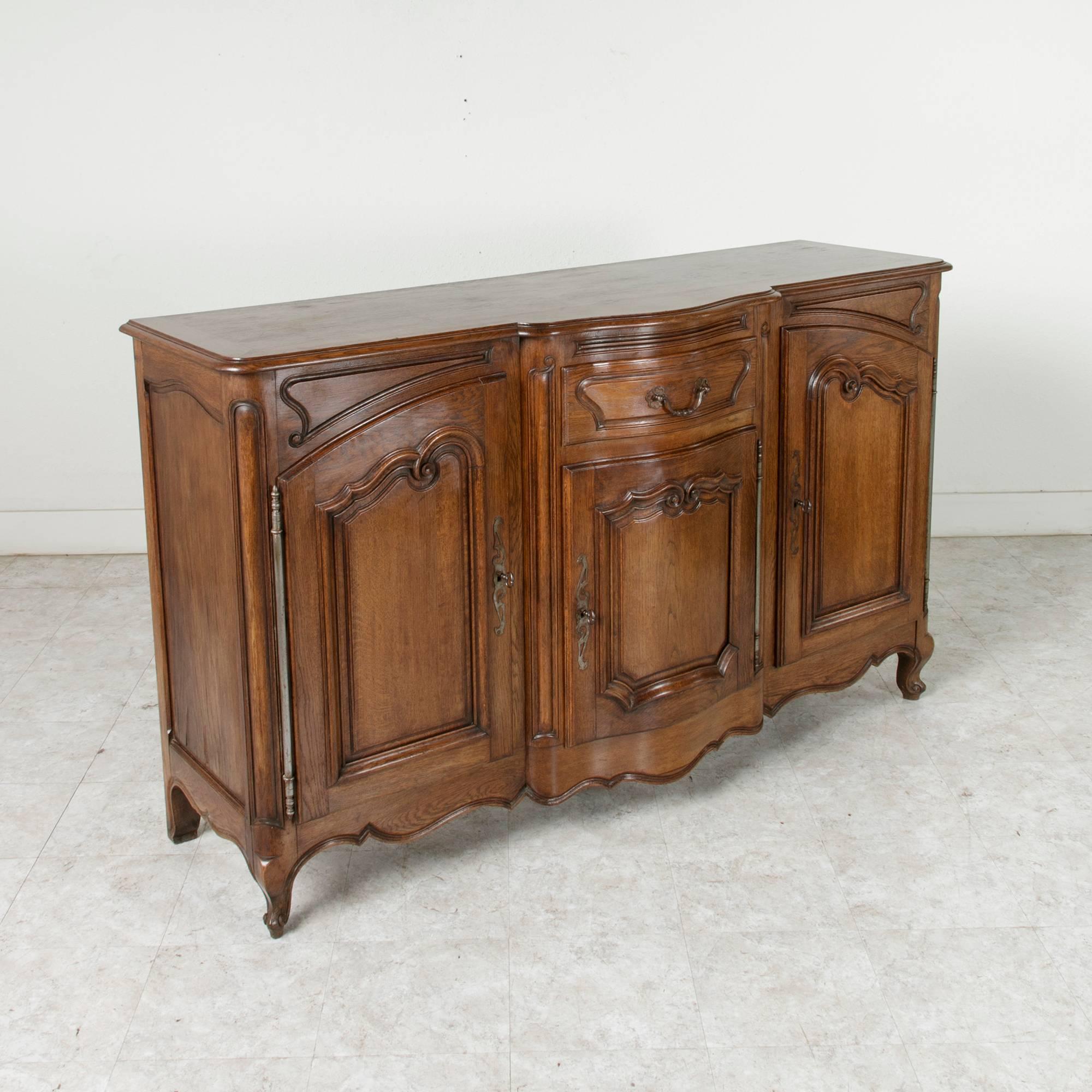This hand-carved Louis XV style oak enfilade or sideboard from the early 20th century features a bow front and solid panel sides with a beveled top. Its bow front contains a single drawer of dovetail construction and a lower door and is flanked by