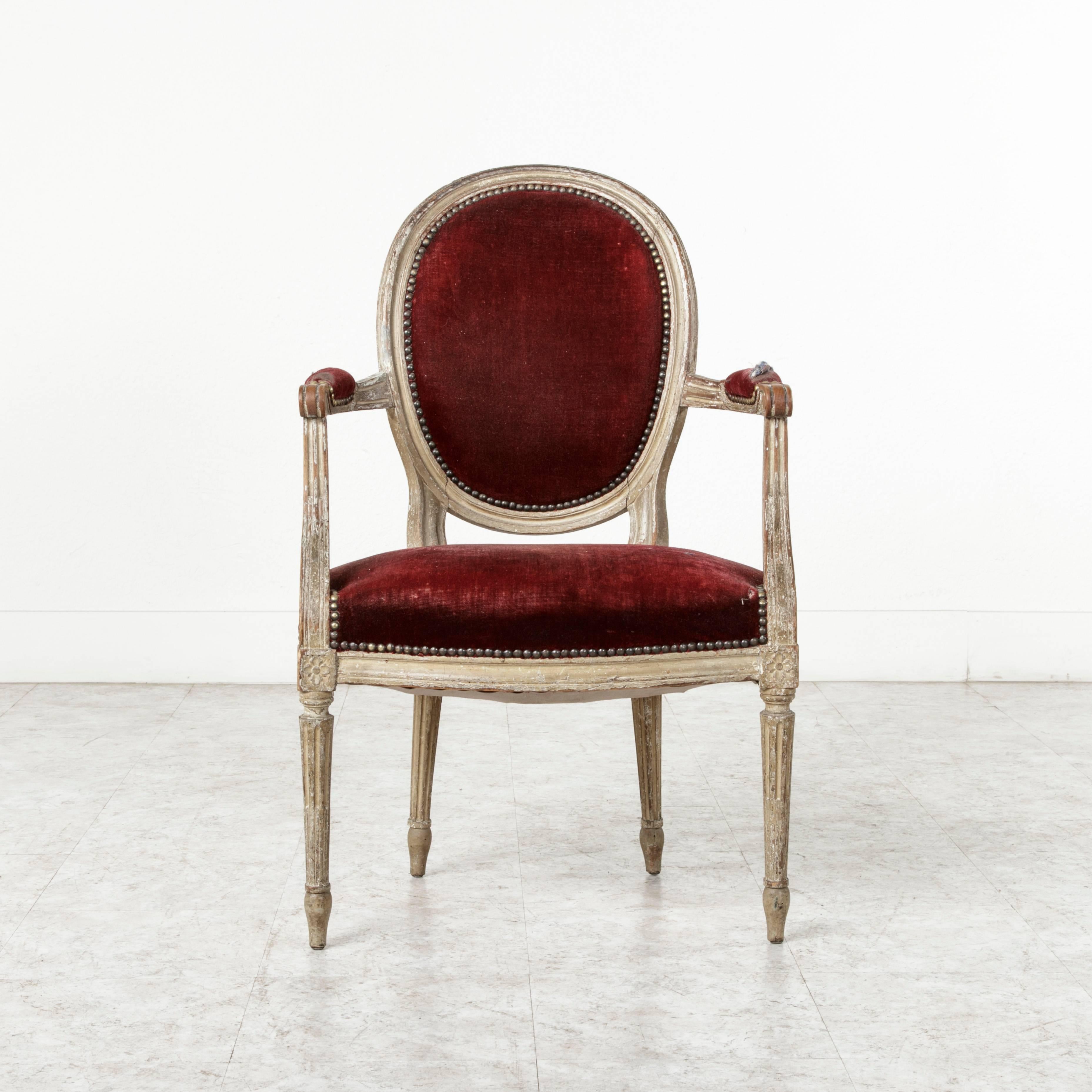 A century of use and vestiges of worn paint have given this Louis XVI cabriolet armchair a patina that only time can create. Its medallion back, cable fluted legs, and rosettes at the die joints are all Classic Louis XVI. Scrolled armrests complete