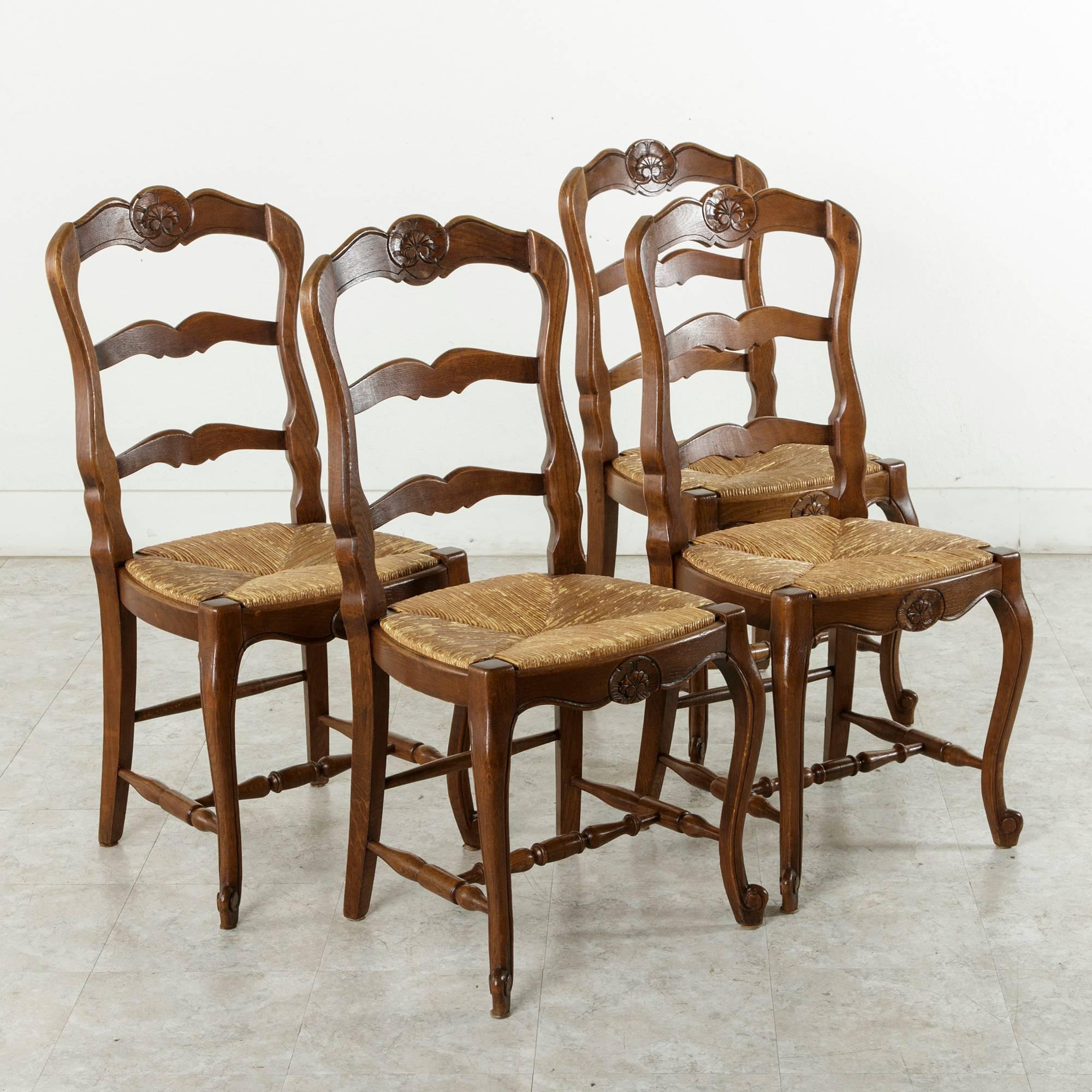 These French oak Louis XV style chairs feature a hand carved shell on both the upper ladderback as well as on the apron. Cabriole legs ending in escargot feet are joined by turned, spooled stretchers on three sides as well as a central stretcher,