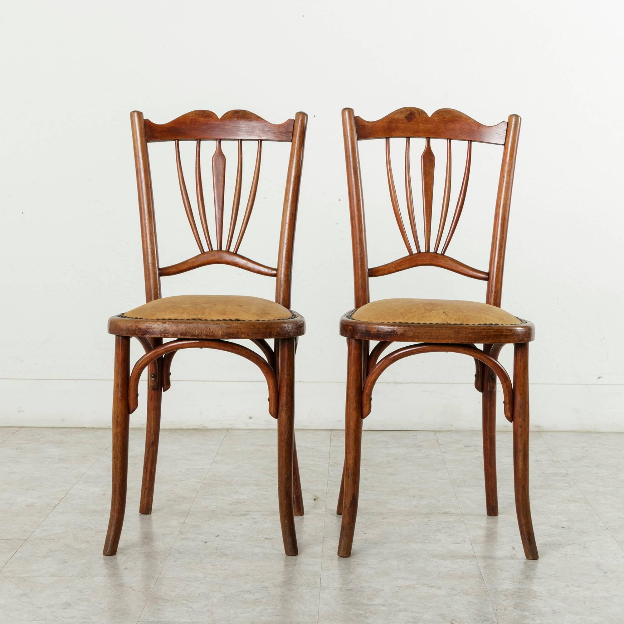 This pair of Art Deco period chairs in the style of Thonet features bentwood frames with their original red toned finish. Their padded cognac leather seats are finished with nailhead trim. Originally used in a French bistro, Paris in the 1920s comes