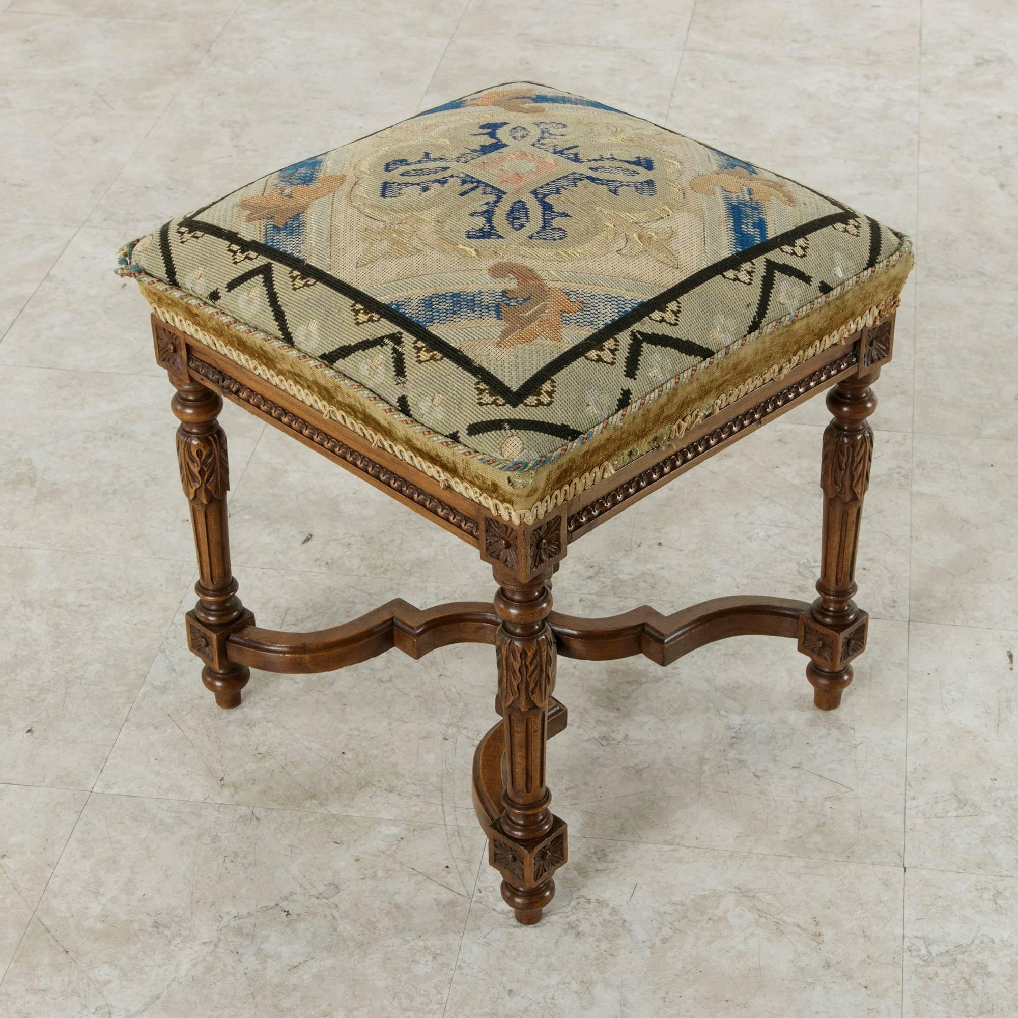 This 19th century hand-carved walnut stool in the Louis XVI style features fluted legs with acanthus leaf carvings at the top and rosettes on the die joints. The elegantly curved X-stretcher with central finial adds stability to this finely detailed