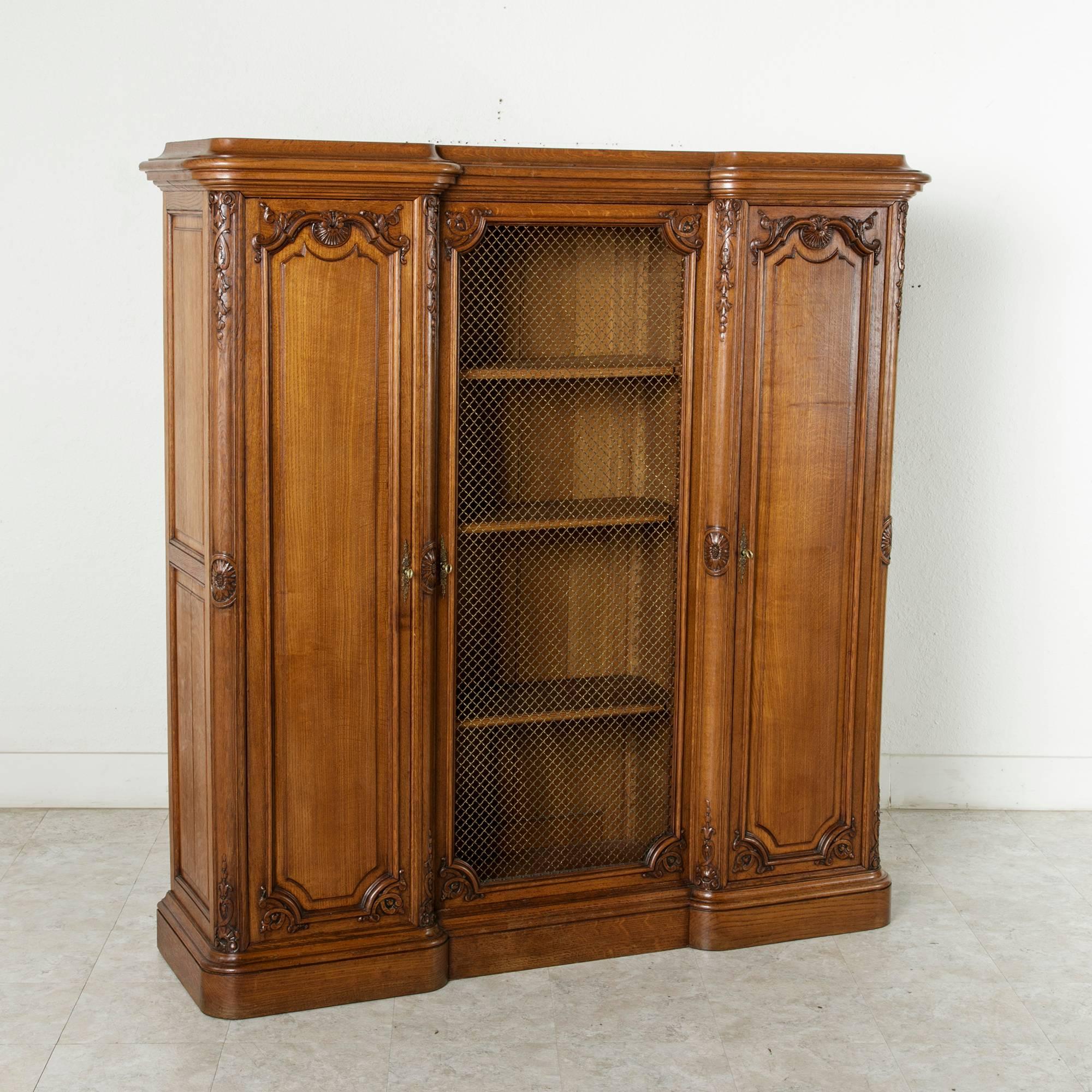 This hand-carved oak French Regency style bibliothèque or bookcase features a central door fitted with bronze wire and flanked by two hand-carved panelled doors. The interior central cabinet has three adjustable shelves, while the flanking cabinets