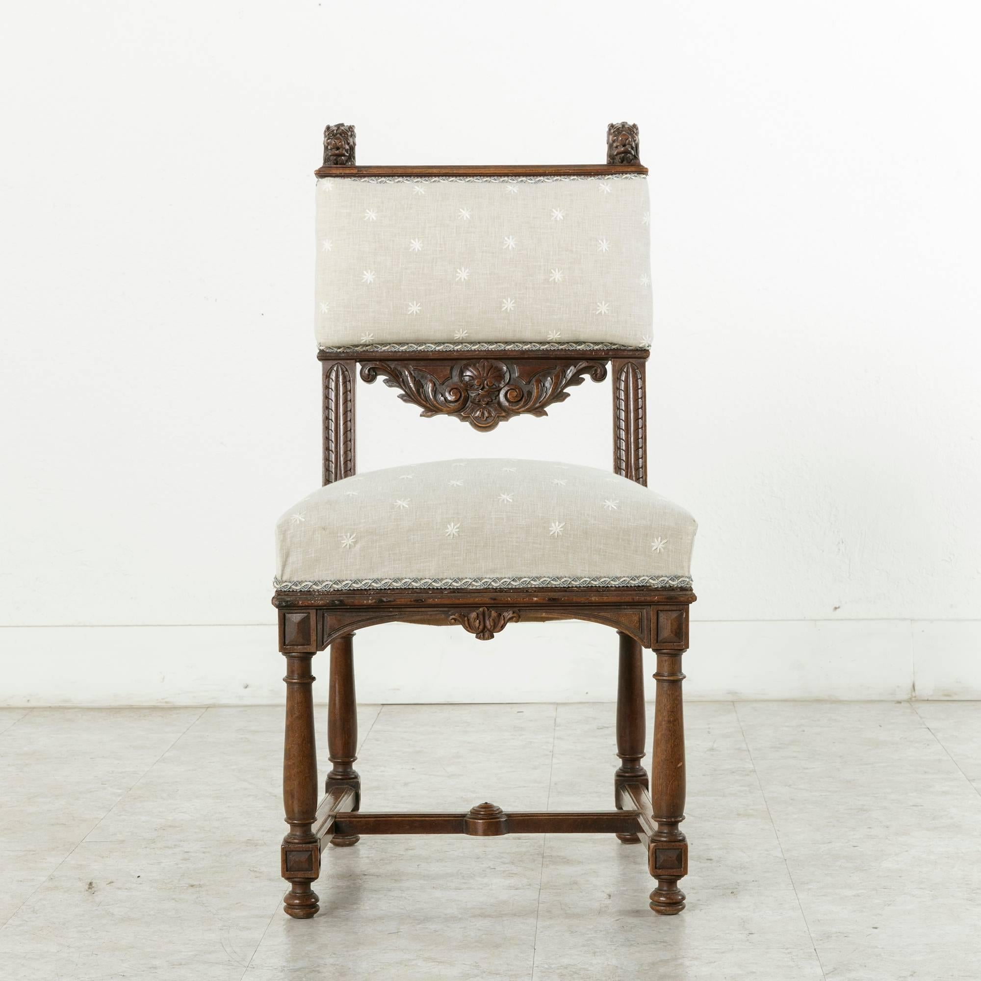 This French hand-carved walnut side chair in the Henri II Renaissance style rests on four tapered column legs joined by an H-stretcher. Its upholstered back is embellished with lion's heads at the top and a deep relief carved scallop with central
