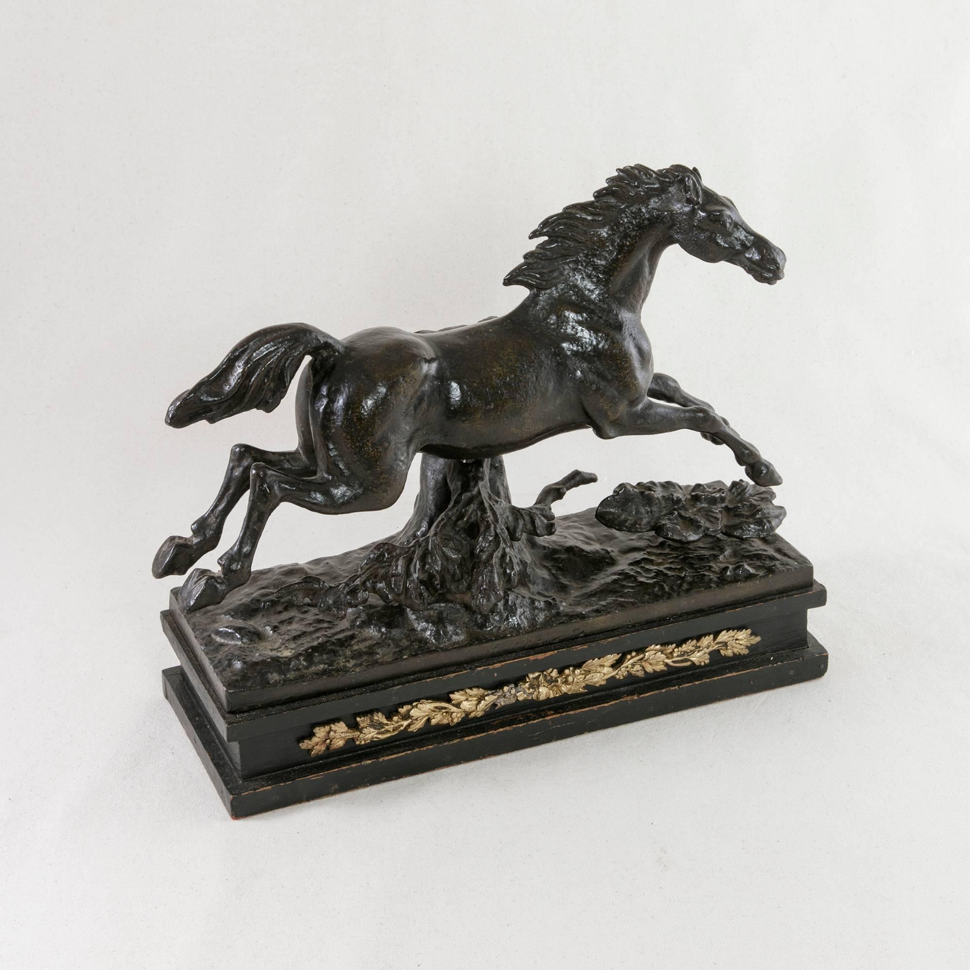 This Napoleon III period cast iron sculpture features a galloping horse near a tree stump surrounded by foliage. It is mounted on a painted black wooden base embellished with a bronze frieze of a central ribbon and bow flanked by branches with