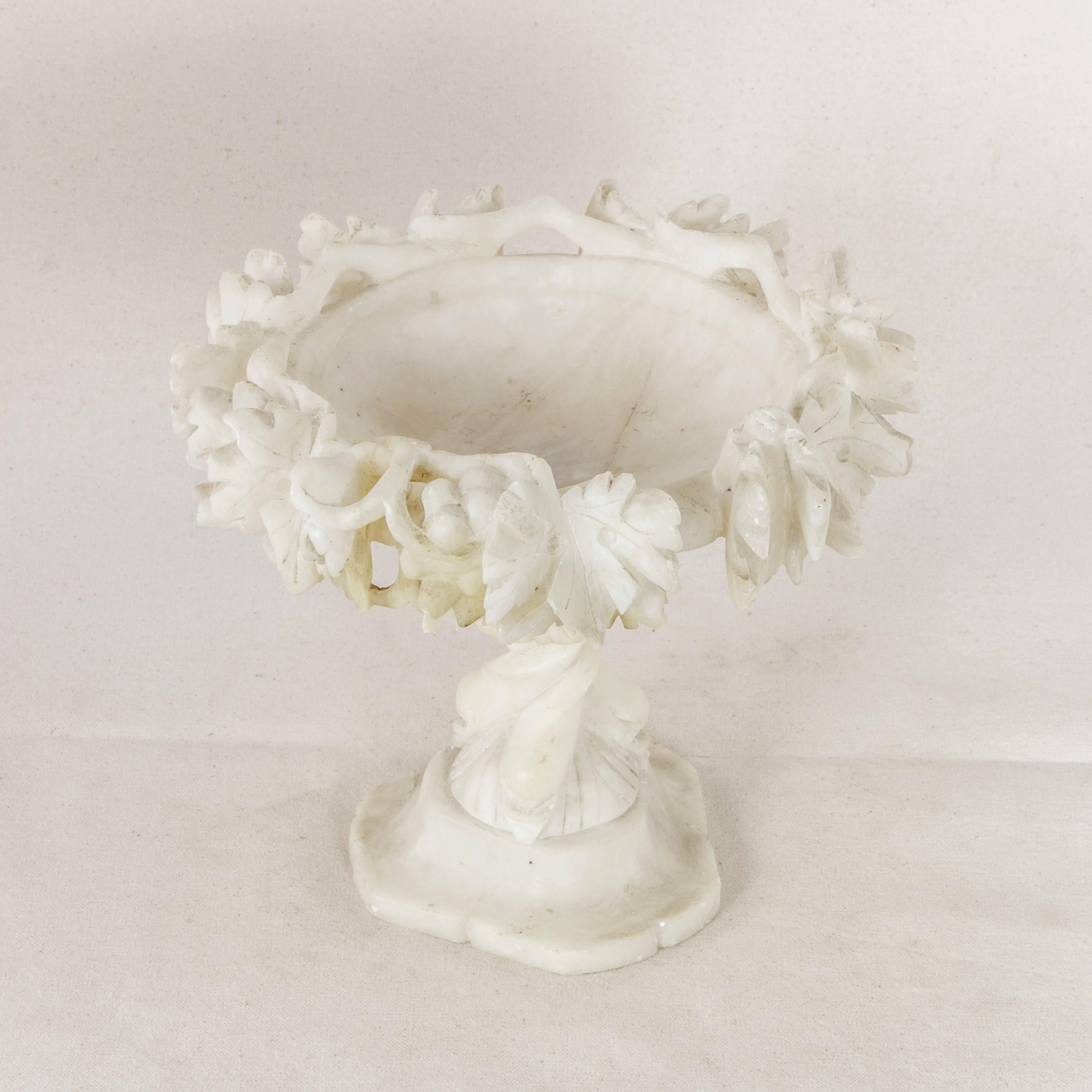 This alabaster compote from the late 19th century features deep relief hand-carved detailing of grape vines and leaves around the rim. Three carved dolphins with intertwined tails form the base and stem. Originally used to serve candies, fruits, or