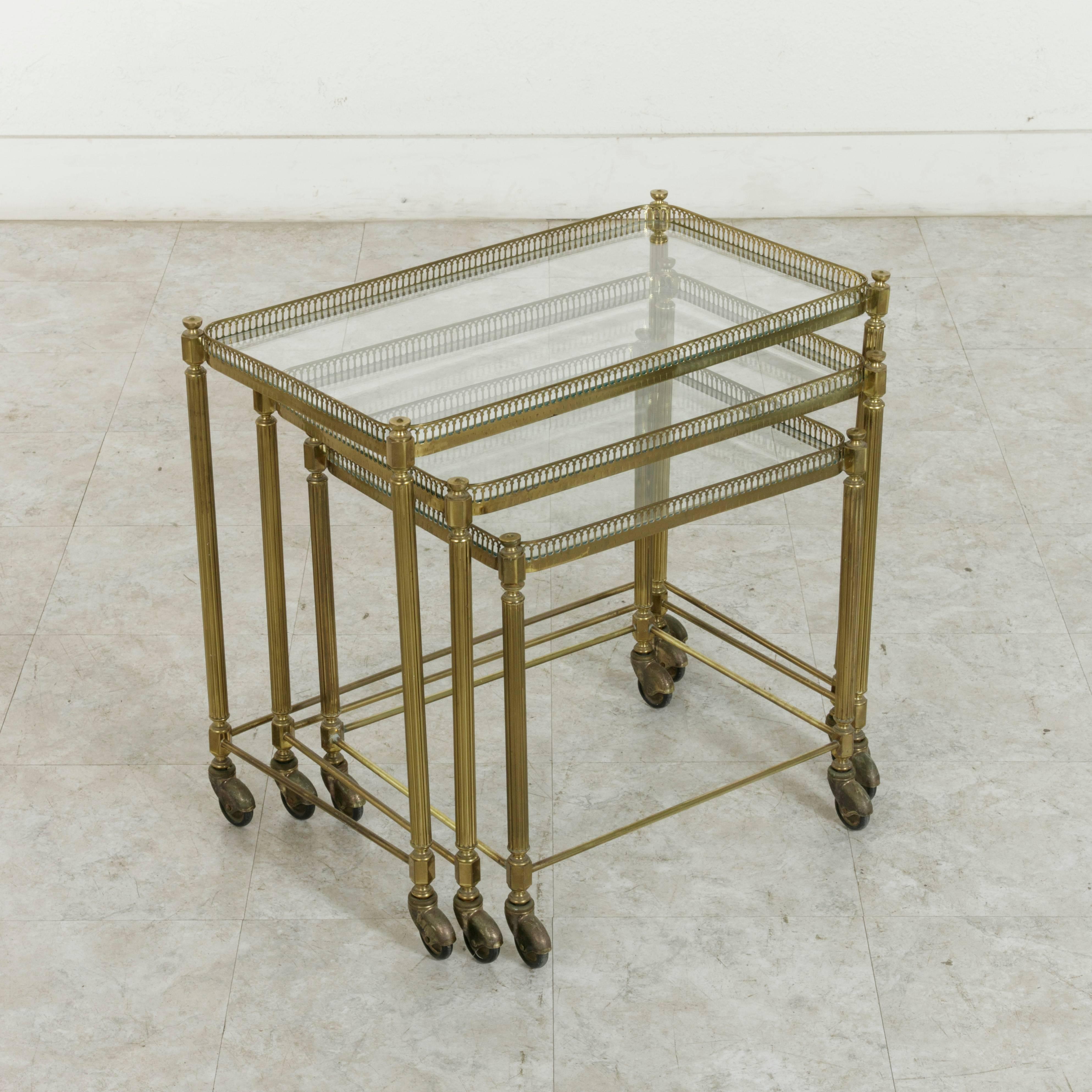 This set of three mid-twentieth century French brass nesting tables features glass tops surrounded by pierced brass galleries. Supported by fluted column legs, they are finished with casters for easy mobility. The largest table measures 24 inches