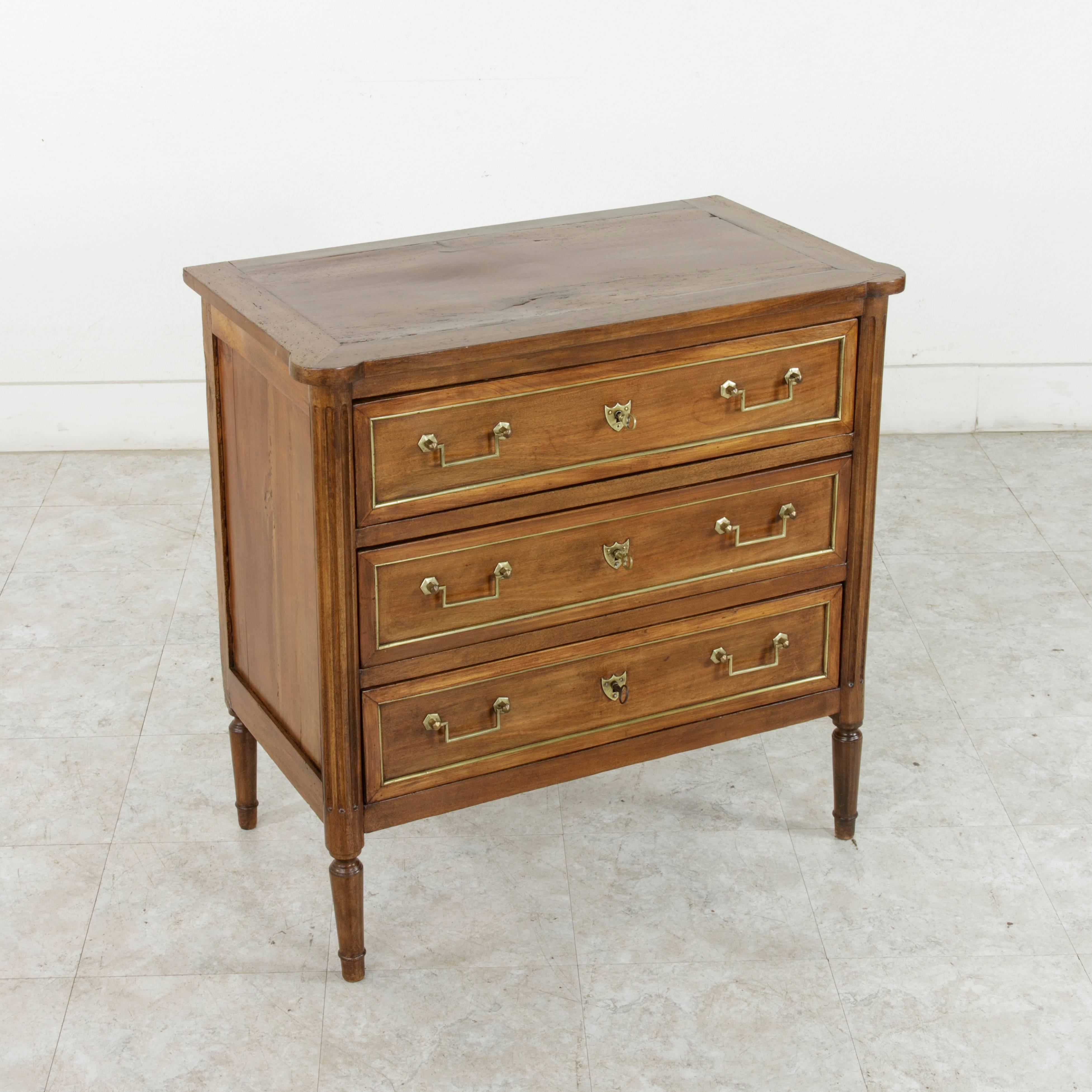 A rare piece from the late 18th century due to its small-scale, this French Louis XVI period walnut commode or chest features fluted corners and rests on tapered legs. The three drawer fronts are detailed with bronze banding and Classic Louis XVI