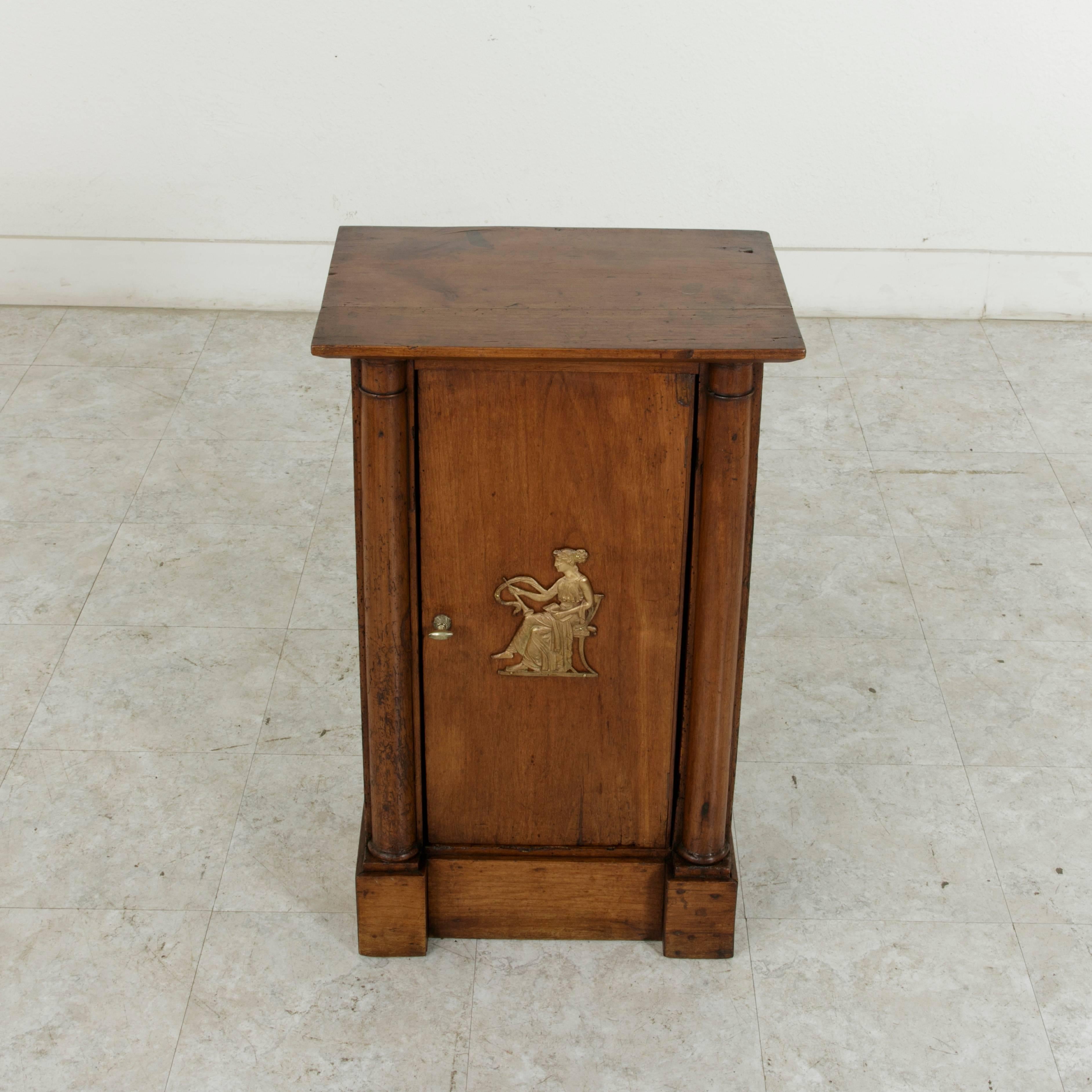 This French Empire period walnut cabinet or nightstand features two half column supports that flank the single door. A brass ornamentation of a classical Greek female figure sitting in a chair adorns the door. In her right hand she holds a flute