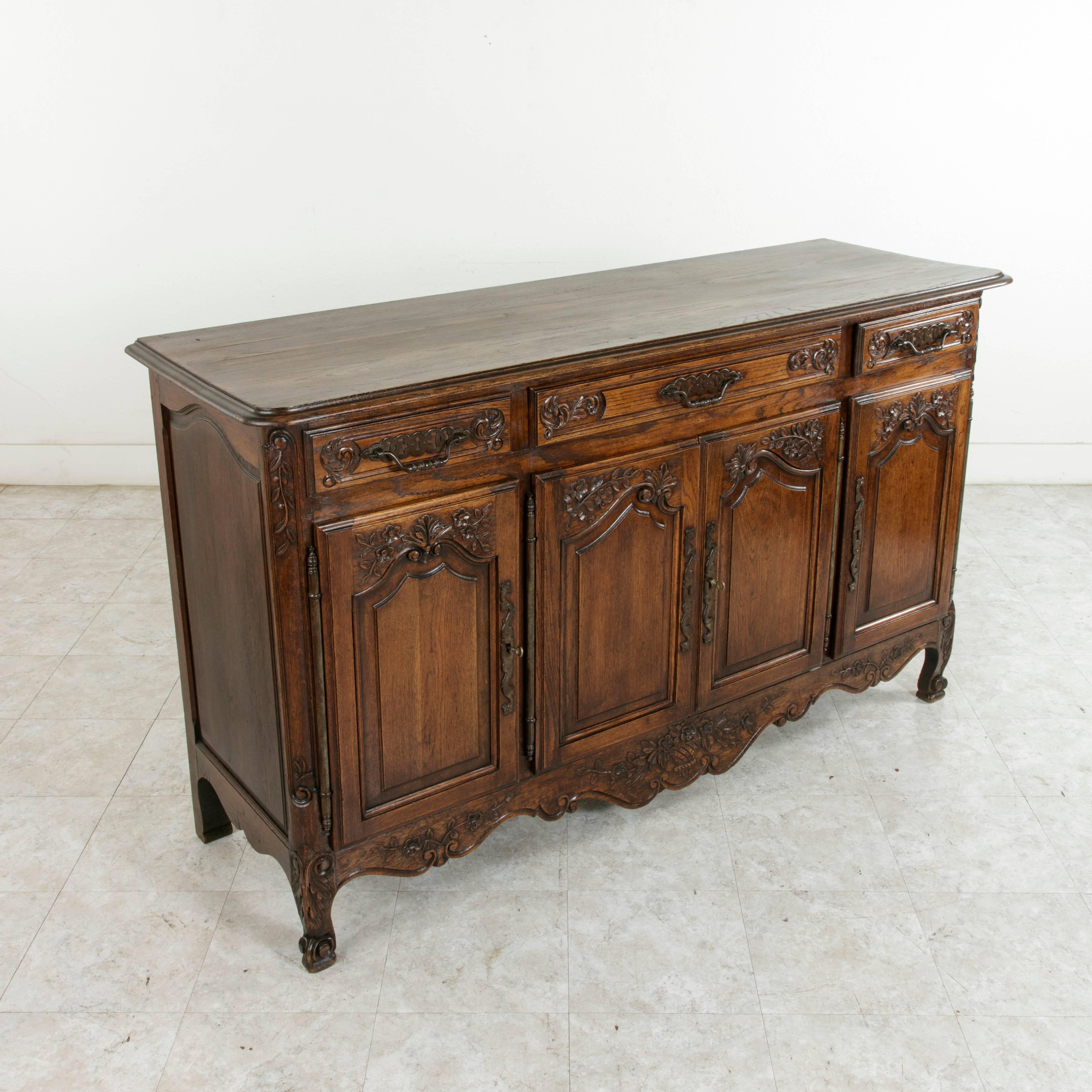 This early 20th century Louis XV style oak enfilade or sideboard from Normandy, France, features paneled sides and doors, and hand-carved motifs of scrolling, leaves, flowers, and shells. A carved basket, the symbol of bounty in the region of