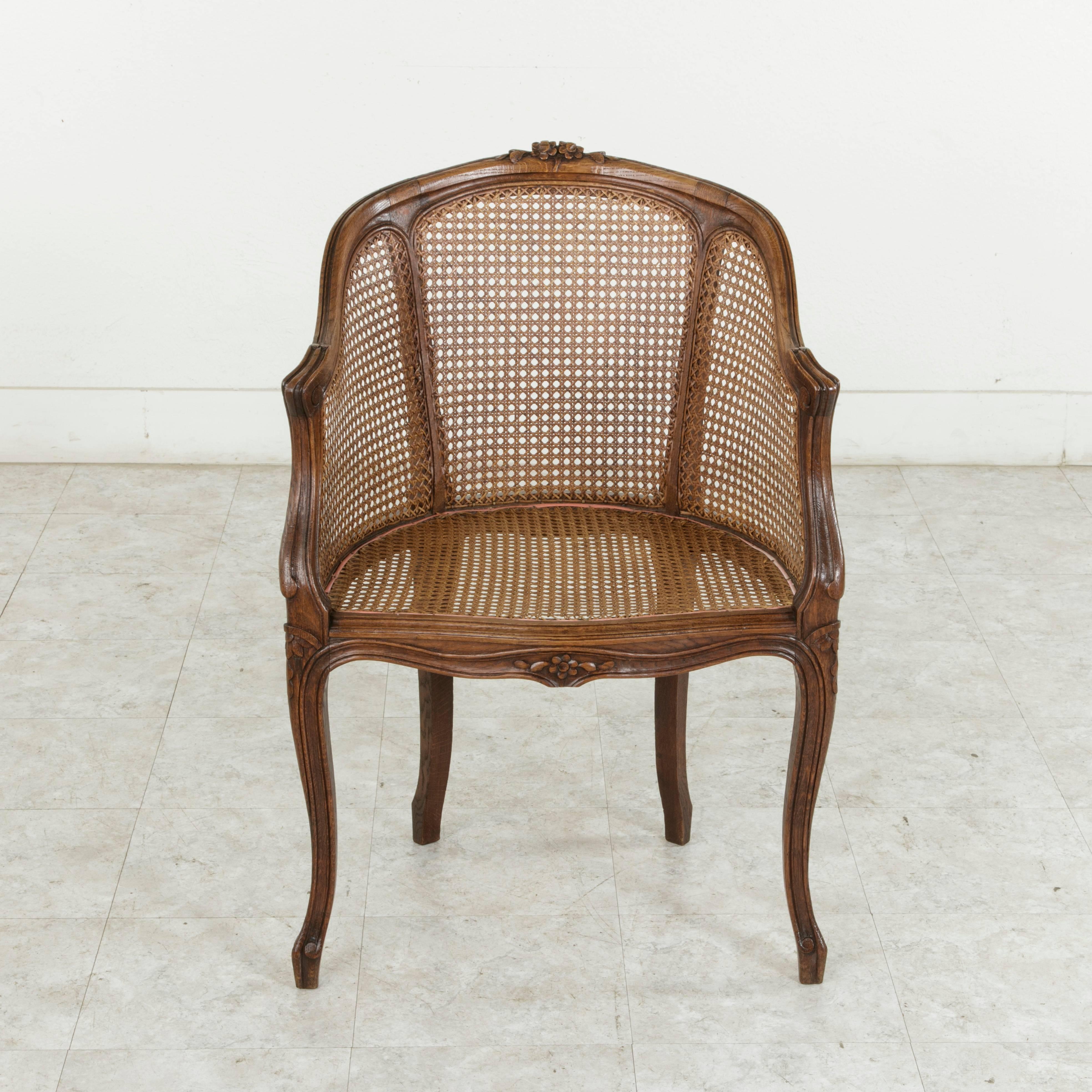 This early 20th century French Louis XV style bergere or armchair features an elegantly curved back with hand-carved details of flowers and leaves at the crown that are repeated on the lower apron. Its caned seat, sides, and back are all in pristine