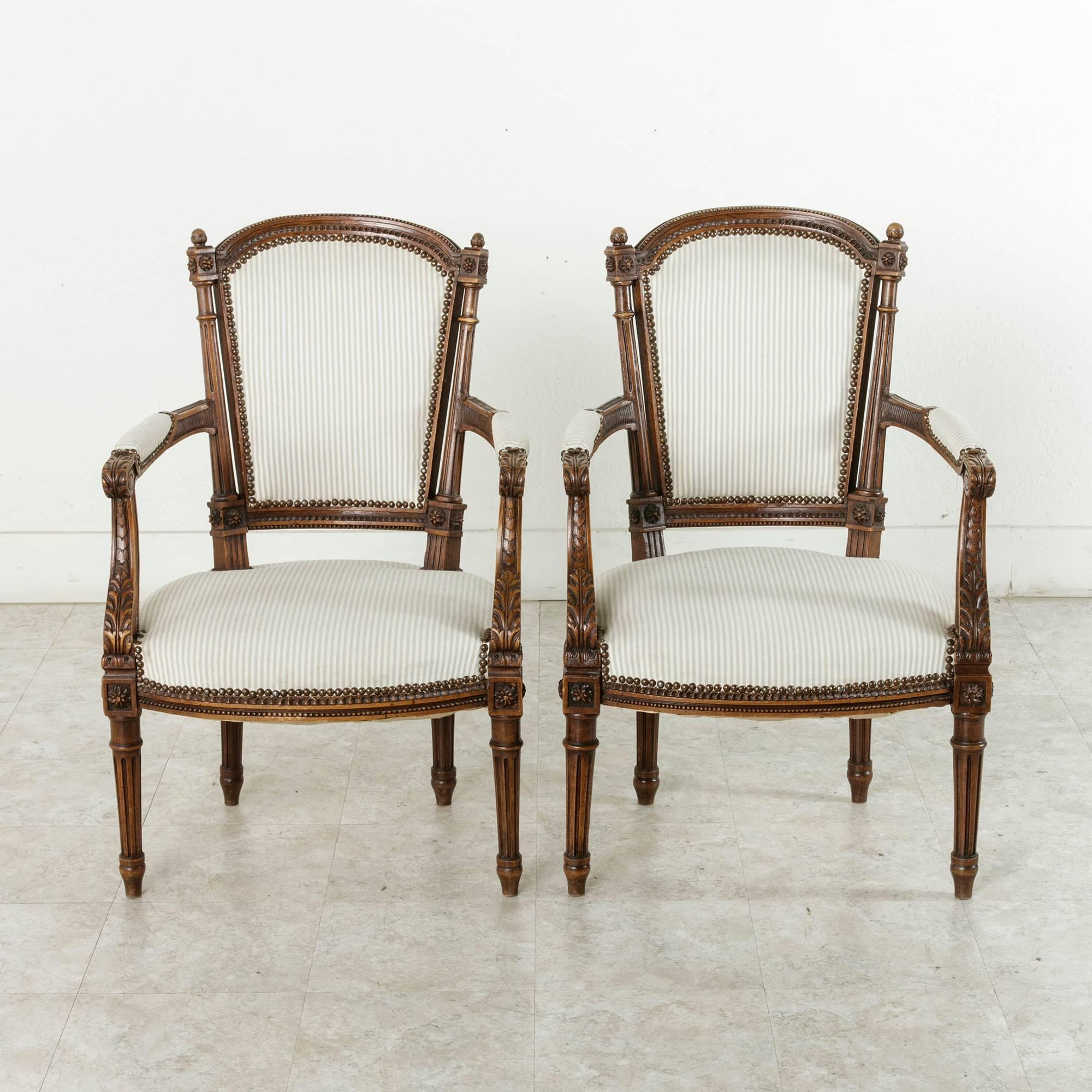 This pair of hand-carved walnut Louis XVI style armchairs from Provence feature classic tapered, fluted legs and high relief rosettes on the die joints.   Carved acanthus leaves adorn the arms. The upholstered chair back is flanked by fluted columns