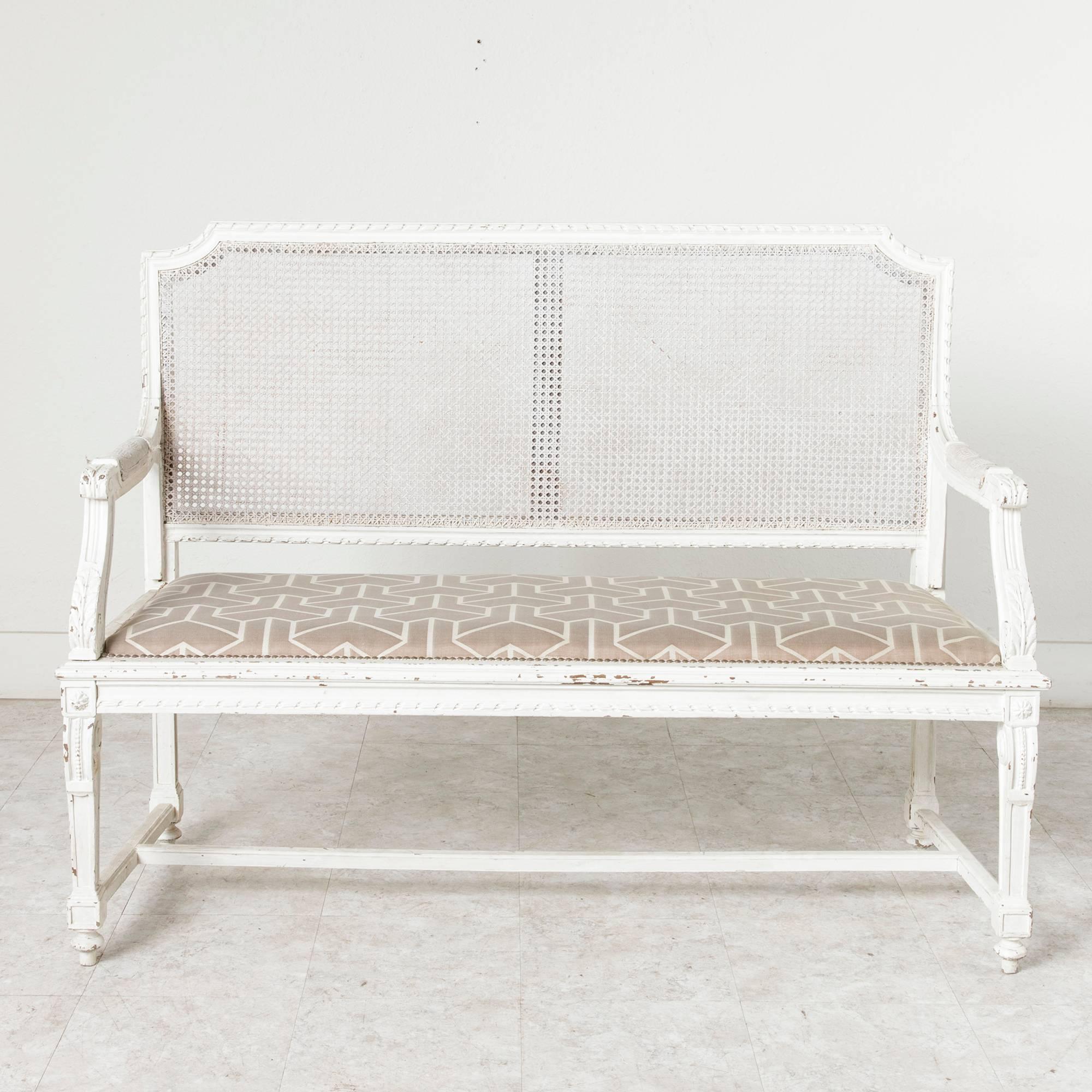 This late 19th century hand-carved Louis XVI style banquette or bench, painted in crisp white, features a caned back and upholstered seat. Perfect for an entryway, garden room, or for sitting on the veranda, circa 1880.