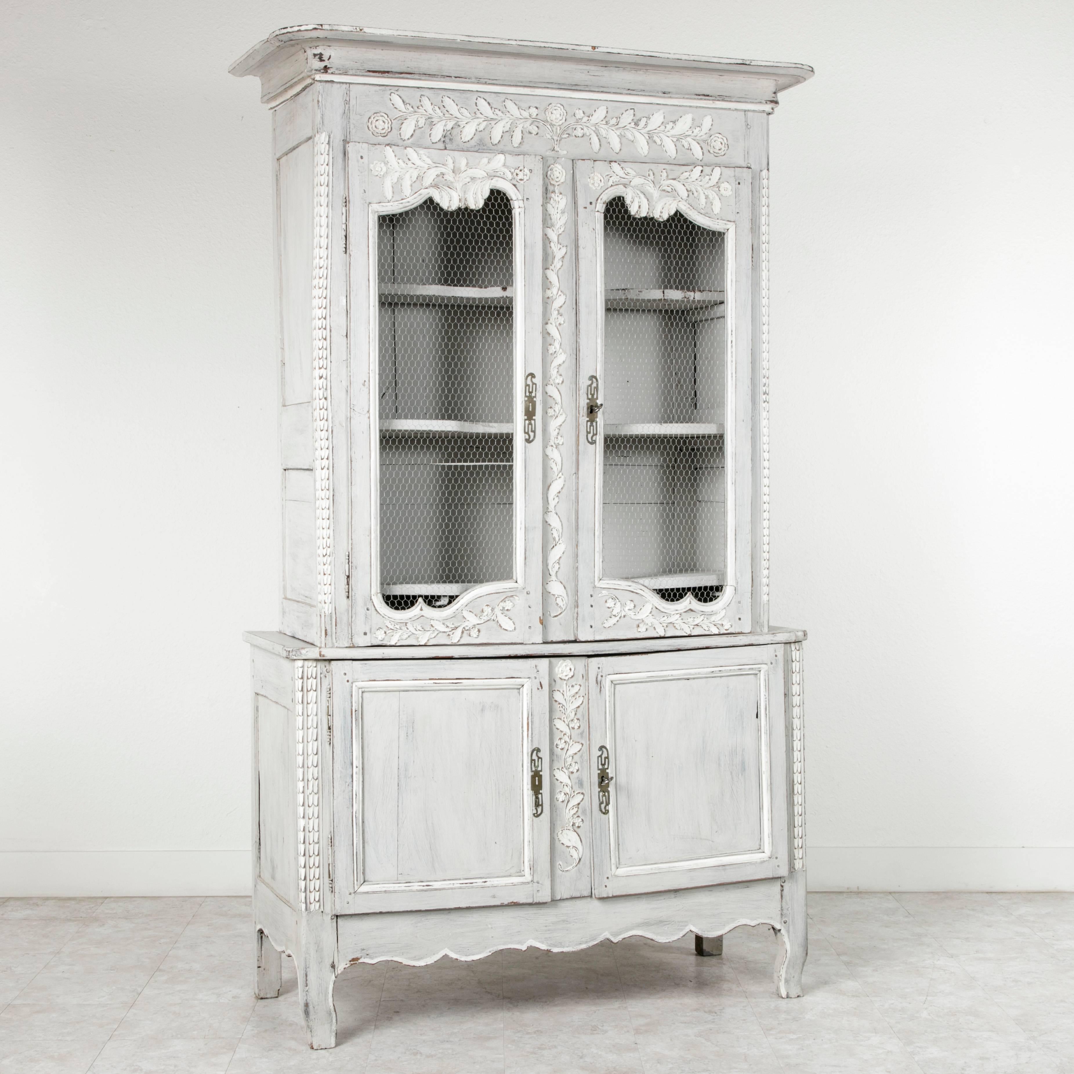 This turn of the century Louis XVI style buffet deux corps is painted in a light hue of blue grey, referred to as Marie Antoinette grey. Its lavish hand carvings of leaves and flowers at the crown and around the upper doors are enhanced by its