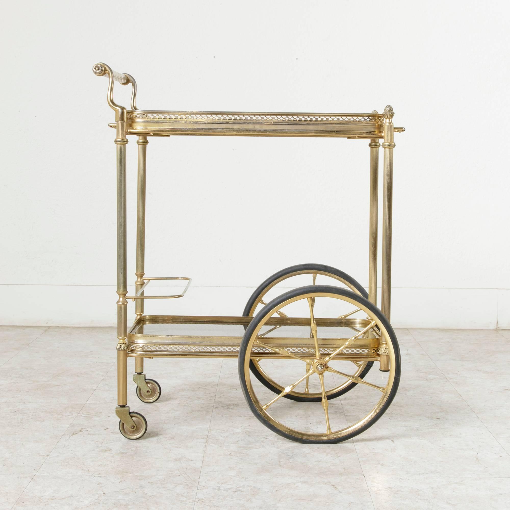 This mid-20th century brass bar cart from France features a brass gallery around each of its smoked glass shelves. Its top shelf is a removable tray, and its four legs are capped with Louis XVI pine cone finials. An elegant piece that is very