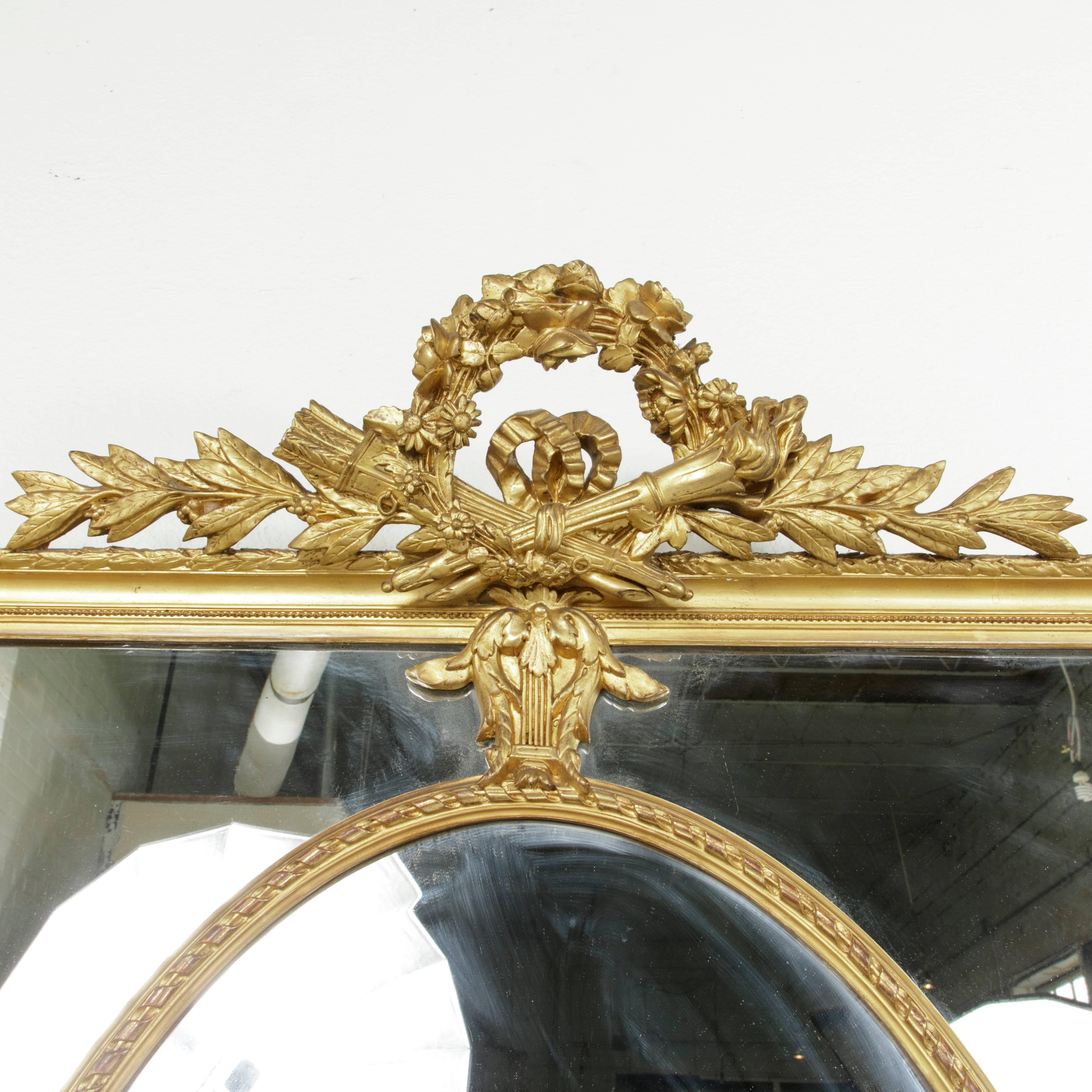 This large late 19th century French giltwood mirror features a classic Louis XVI motif of crossed torch and quiver of arrows with a central floral crown at the top. Hand-carved lyres connect the outer frame to the inset giltwood oval that surrounds
