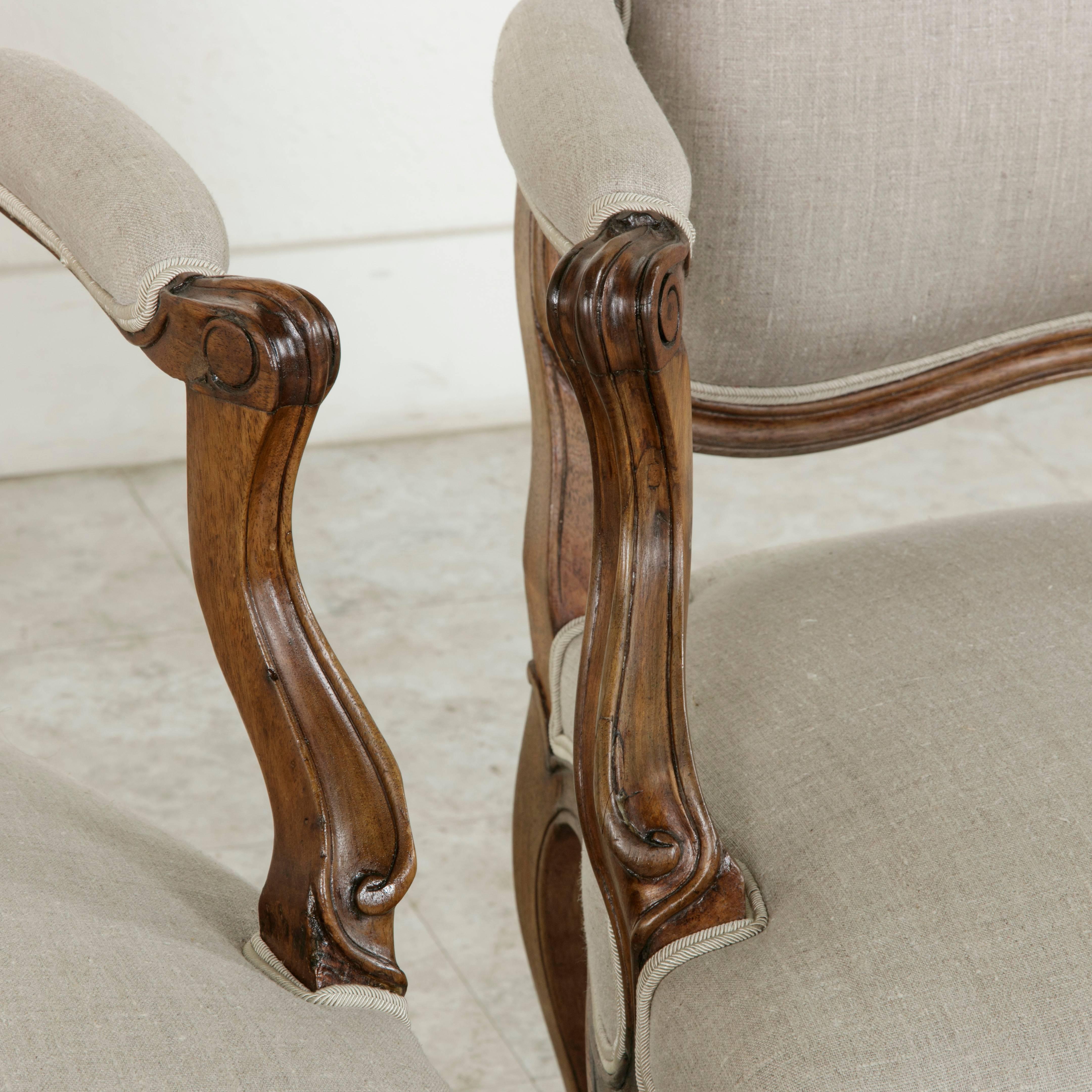 This pair of nineteenth century hand carved walnut armchairs is presented in the classic French Louis XV style. With gently curved cabriole legs and hand carved scrolled armrests, this fine pair includes details of carved flowers at the corners of
