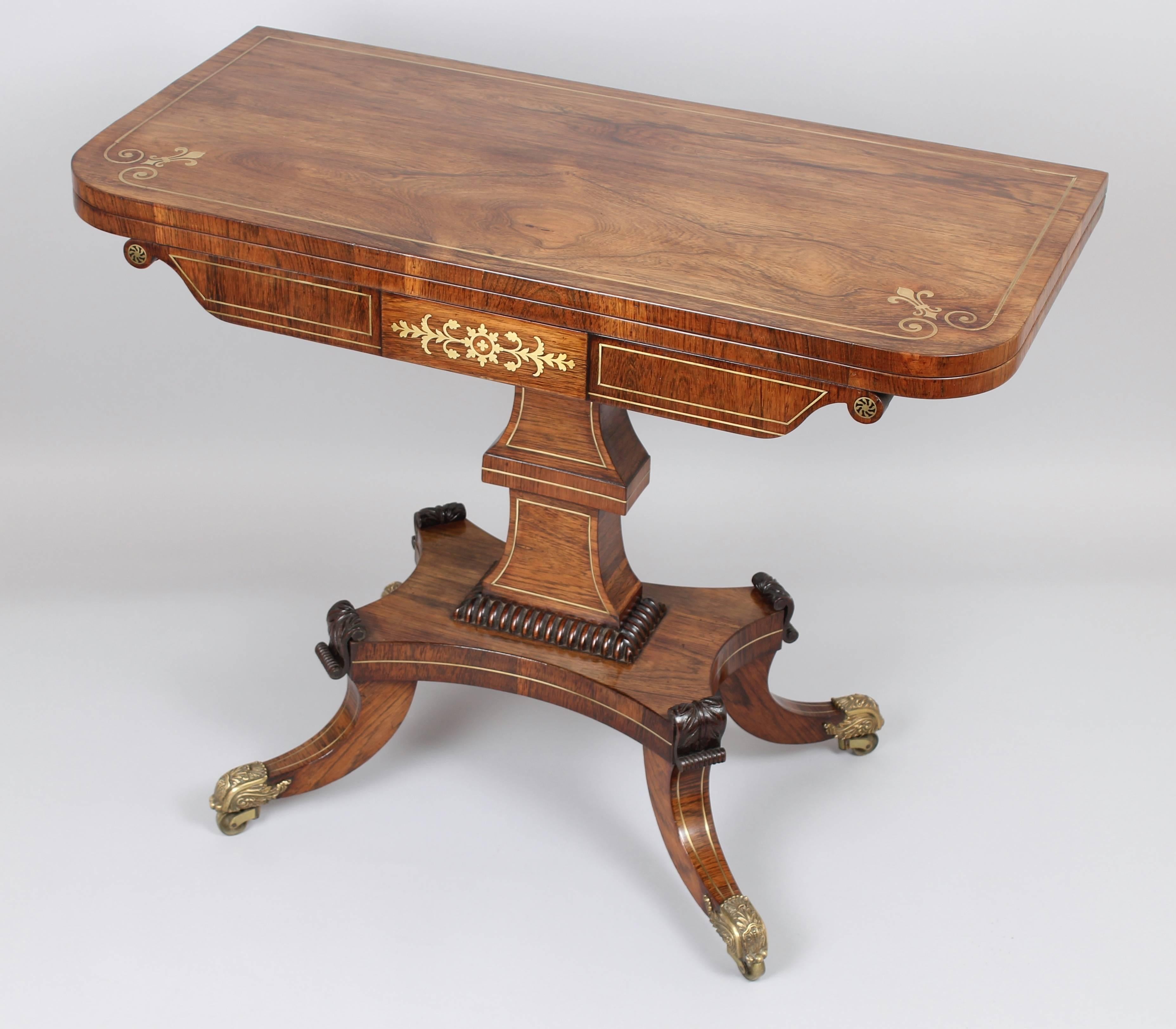 Fine quality Regency rosewood and brass inlaid card-table of mellow golden colour; the hinged and swivelling top inlaid with brass lines and fleur-de-lys; on a shaped frieze with cut-brass scrollwork and paterae; on an elaborate double-blocked shaft