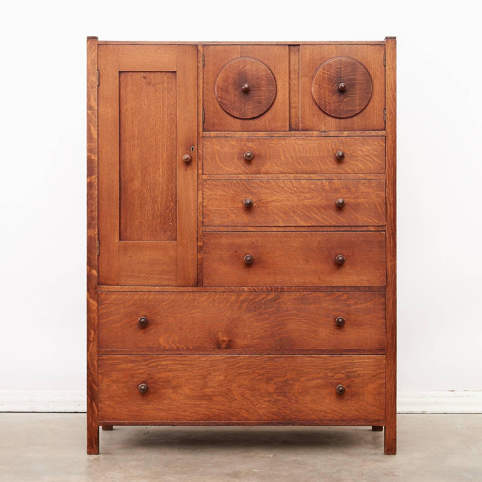 Designed by Ambrose Heal, circa 1902, a solid oak, Arts and Crafts chest of drawers with a pair of 'owl's eye' cabinet doors and a large cabinet door with later shelves, all constructed of solid quarter sawn oak.