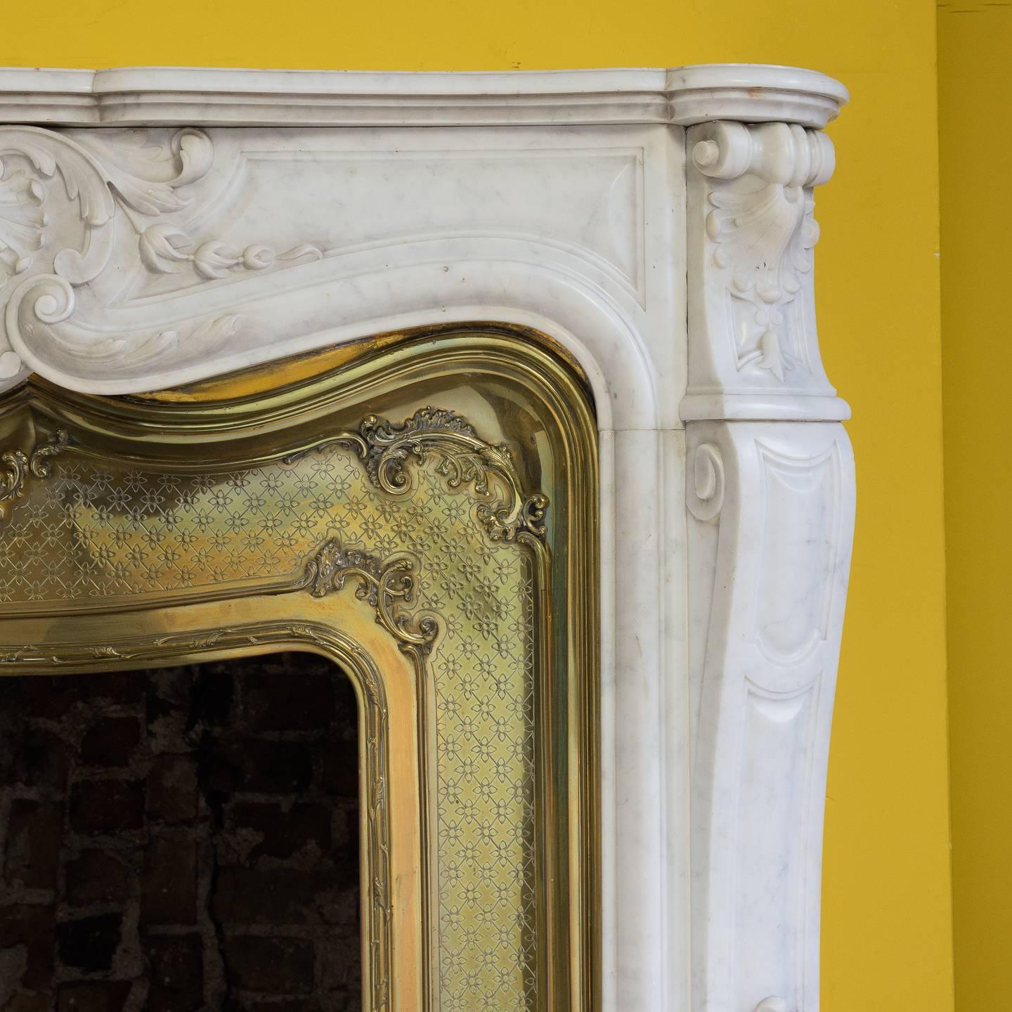 A Louis XV style Carrara marble chimneypiece, 19th century, the shaped and moulded shelf above frieze centered by stylized scallop shell flanked by acanthus flourishes and trails of flowers, the canted jambs with conforming decoration, with brass