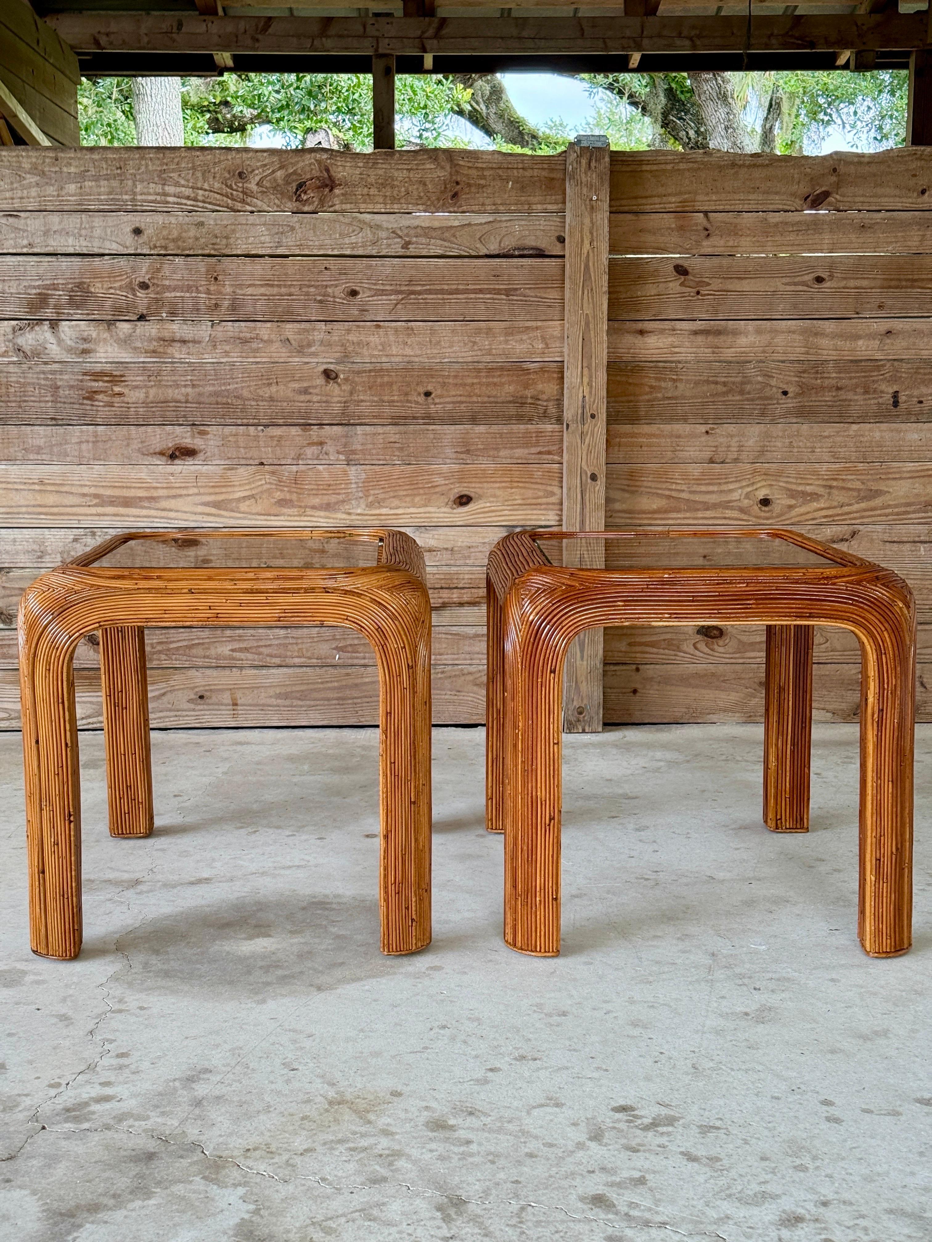A stylish pair of Mid-20th Century pencil reed rattan side tables showcasing bold lines, curved edges, and timeless coastal appeal. Each table features a handcrafted waterfall silhouette with a meticulous pencil reed wrap and newly added glass tops