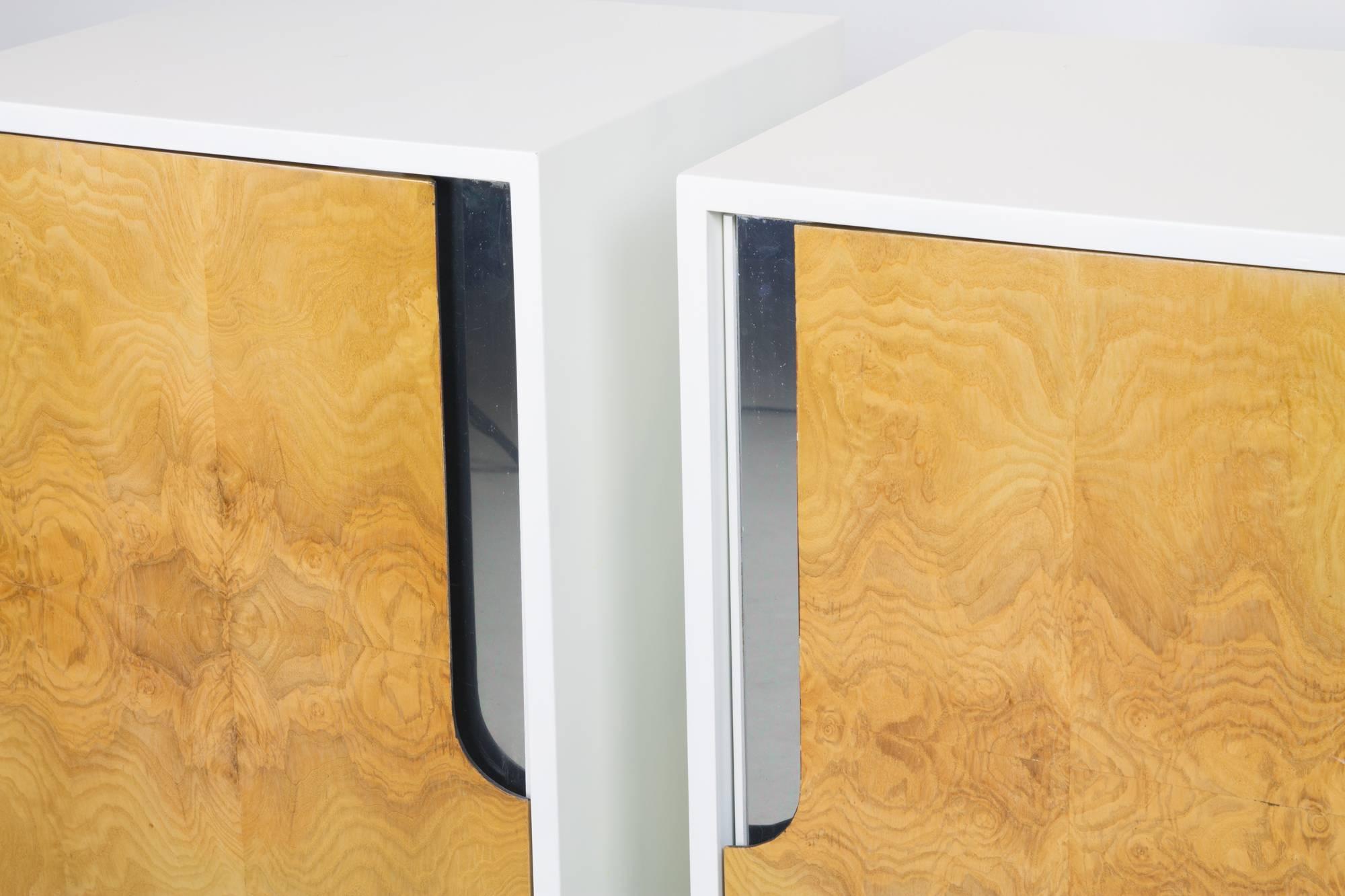 A pair of nightstands by Milo Baughman in white, each with one interior shelf. The doors feature figural bookmatched burl veneer and slivers of mirrored surface behind the pulls.