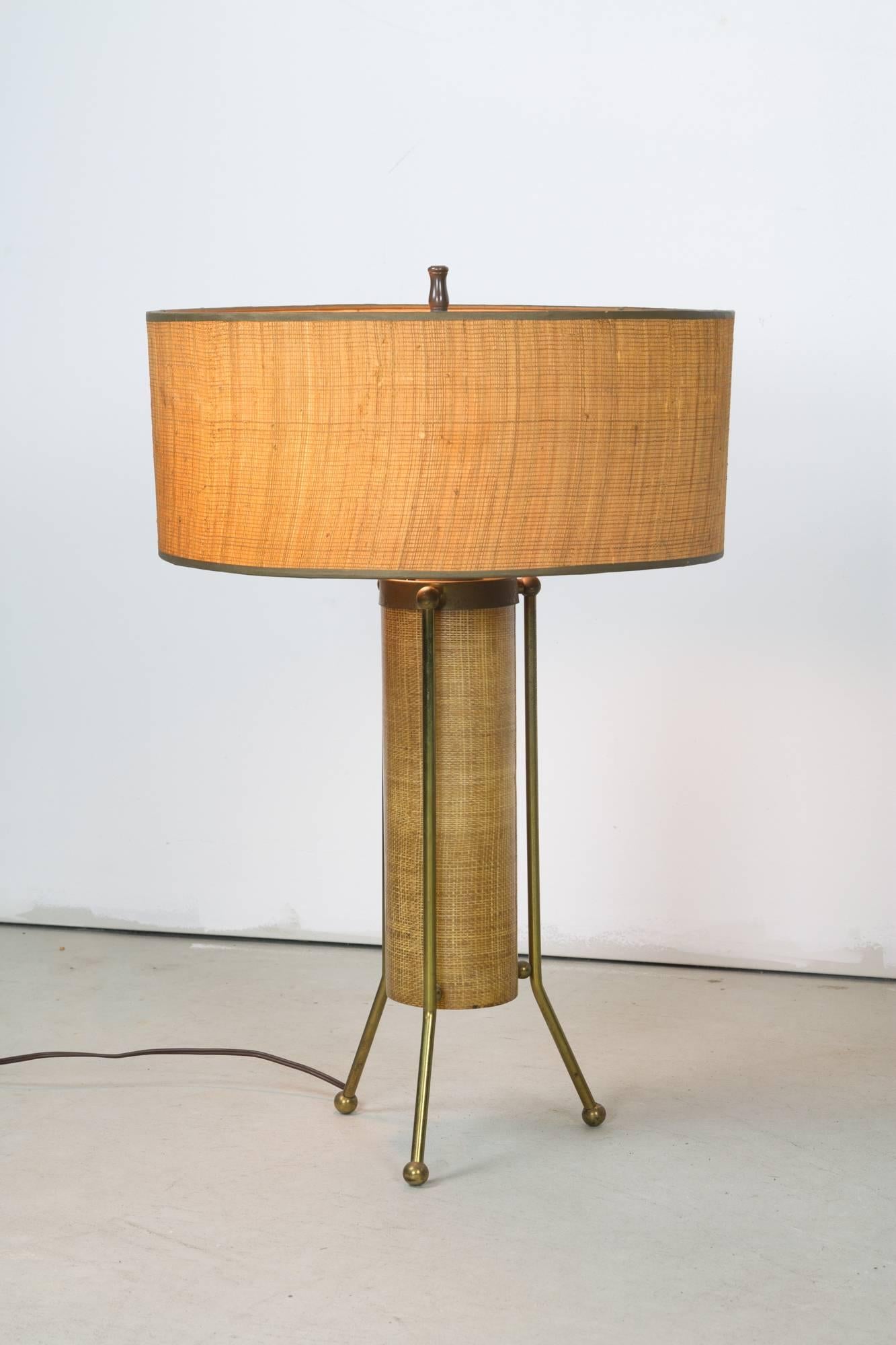 An atomic table lamp featuring a reed shade with stitched fiberglass diffuser and cylindrical fiberglass body-shade producing downlight, supported by an outer brass rod frame and three legged base on ball feet. Light can be cast from the main shade