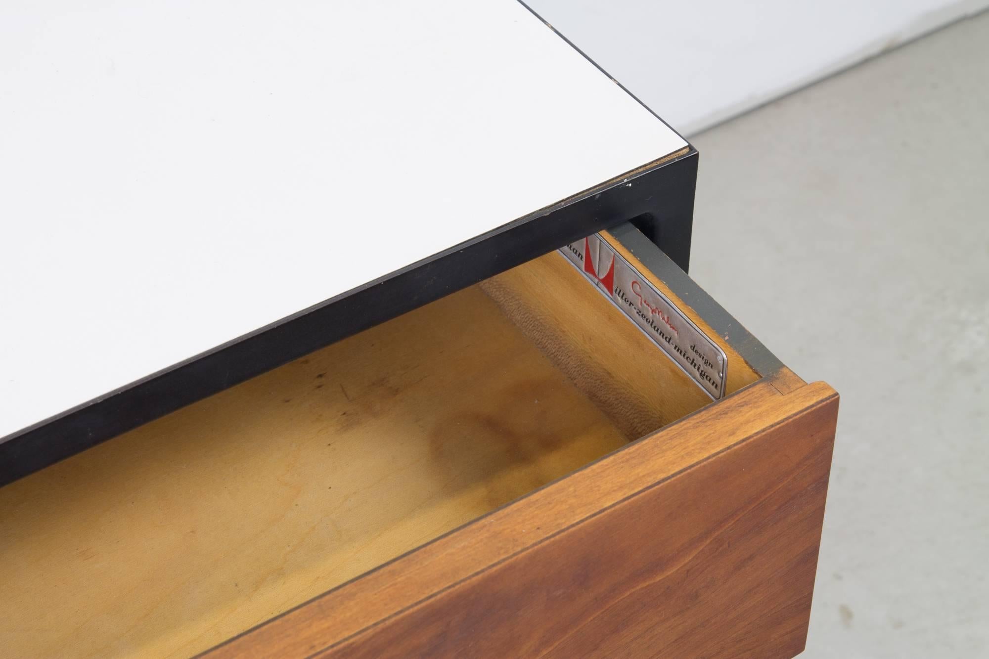 A compact nightstand by George Nelson as part of the steel frame line for Herman Miller. This nightstand features an external frame of blackened steel, with three walnut front drawers, a lower walnut shelf, chromed pulls and a white laminate top.