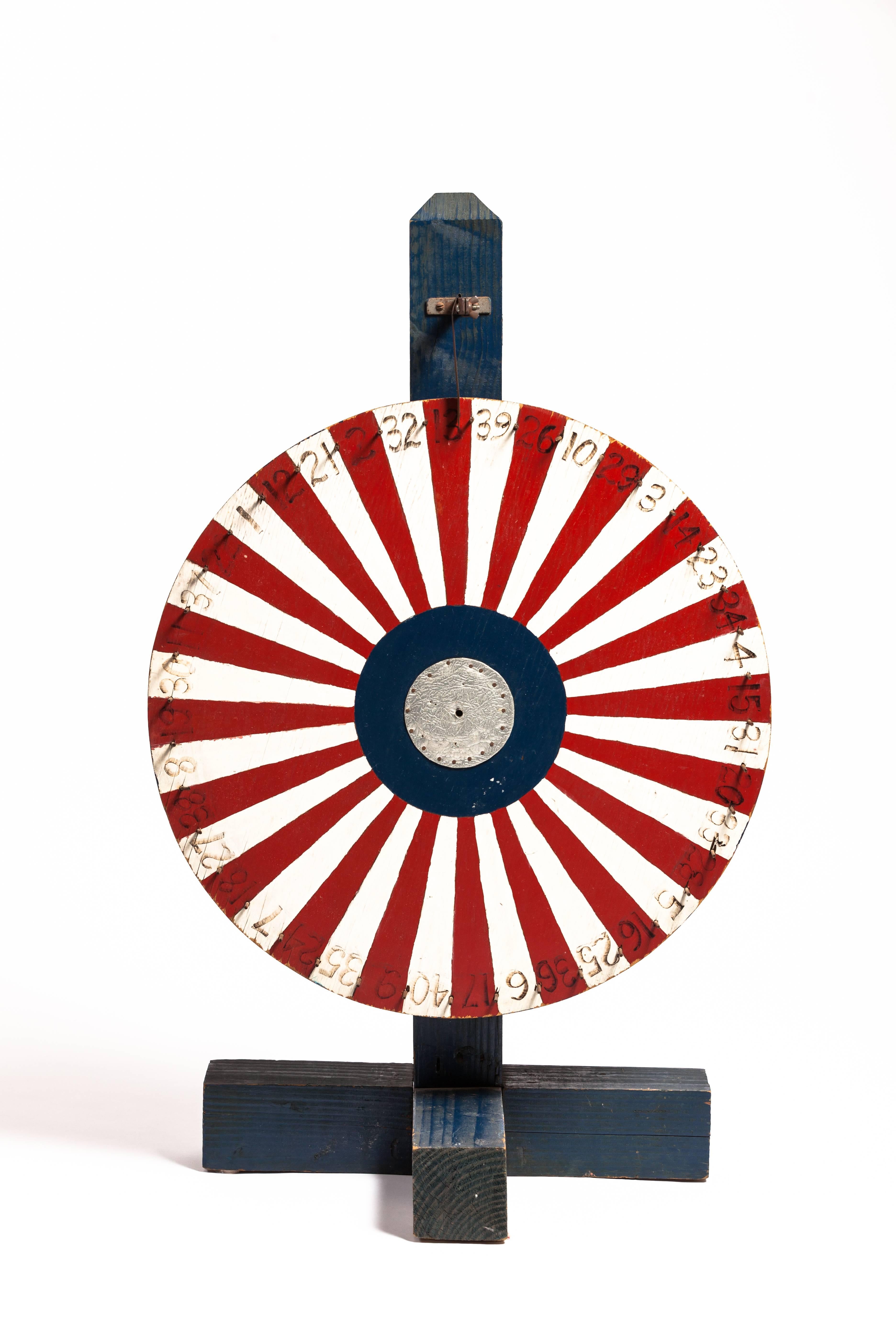 A beautiful Folk Art wooden gaming wheel on a wooden stand or base. The wheel is hand-painted in red white and blue with a silver painted leather center. 

Measures: Base is 20