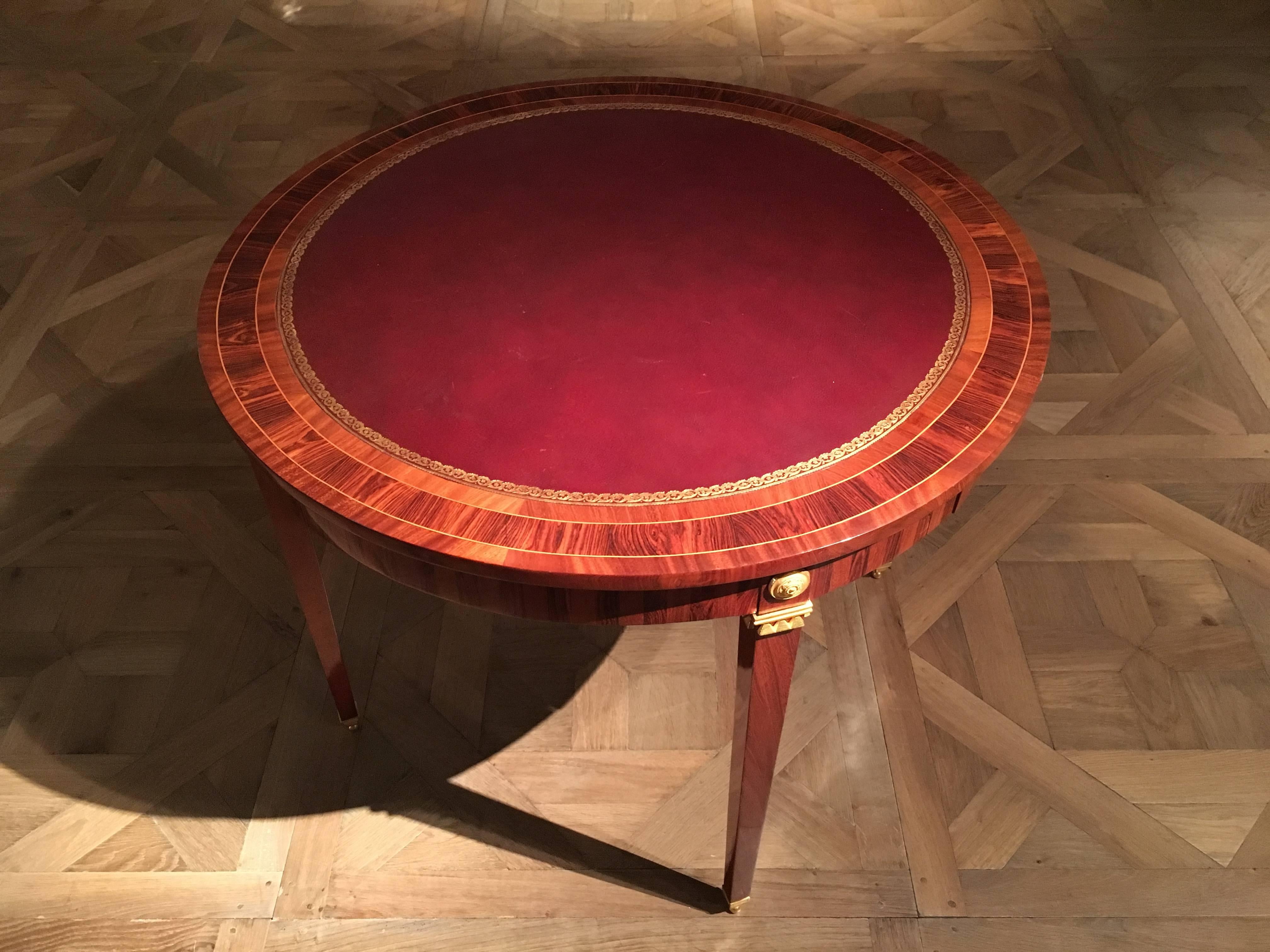 Circular red leather lined top with tulipwood border, the freeze with four small drawers with leather lined sliding cover, raised on four square tapering legs, headed by ormolu rosettes and mounts, standing on ormolu ball feet.