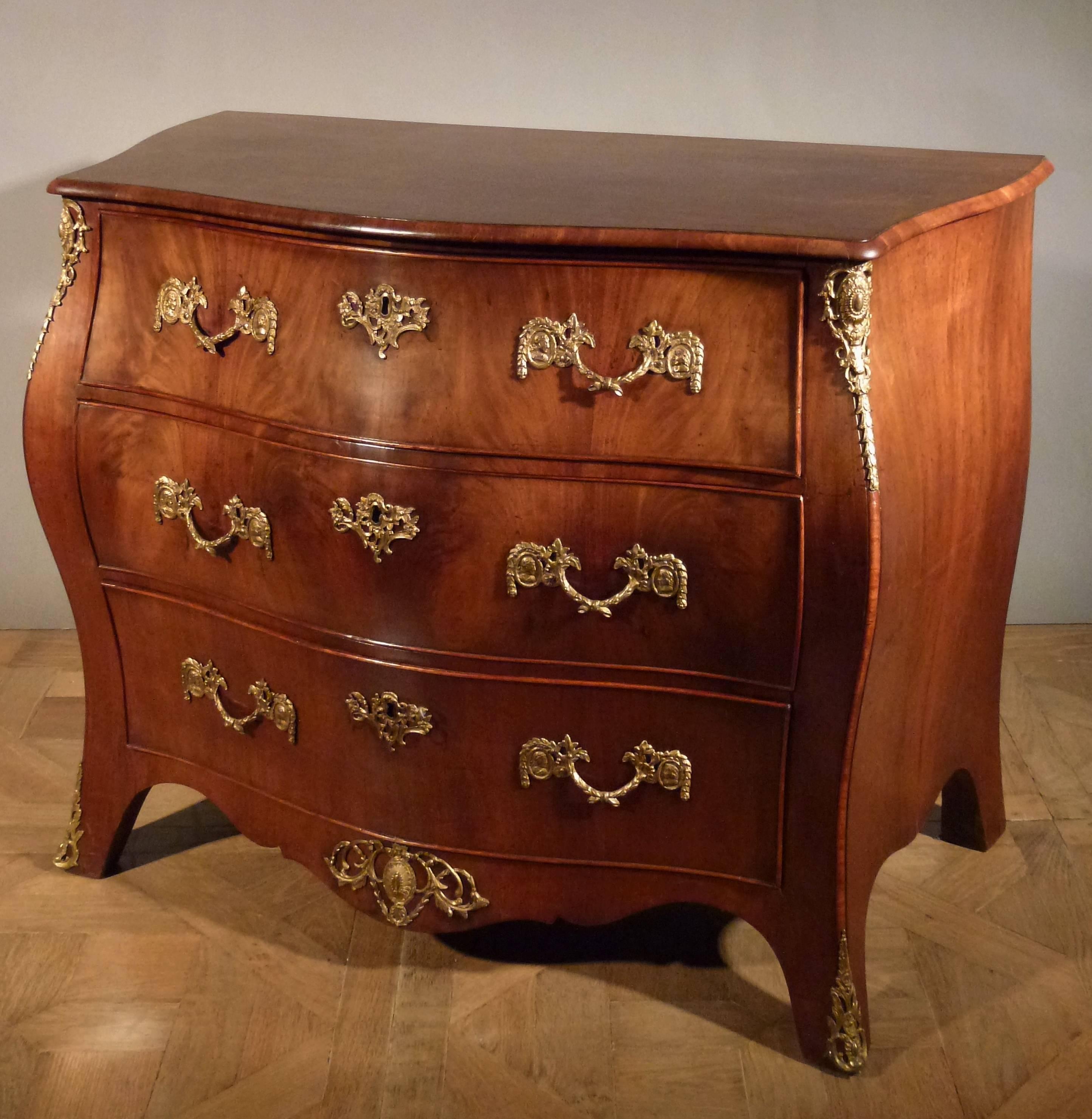 Veneered with mahogany.
The serpentine front with three long drawers, with shaped apron, on splayed feet with pierced sabots, mounted with ormolu handles.