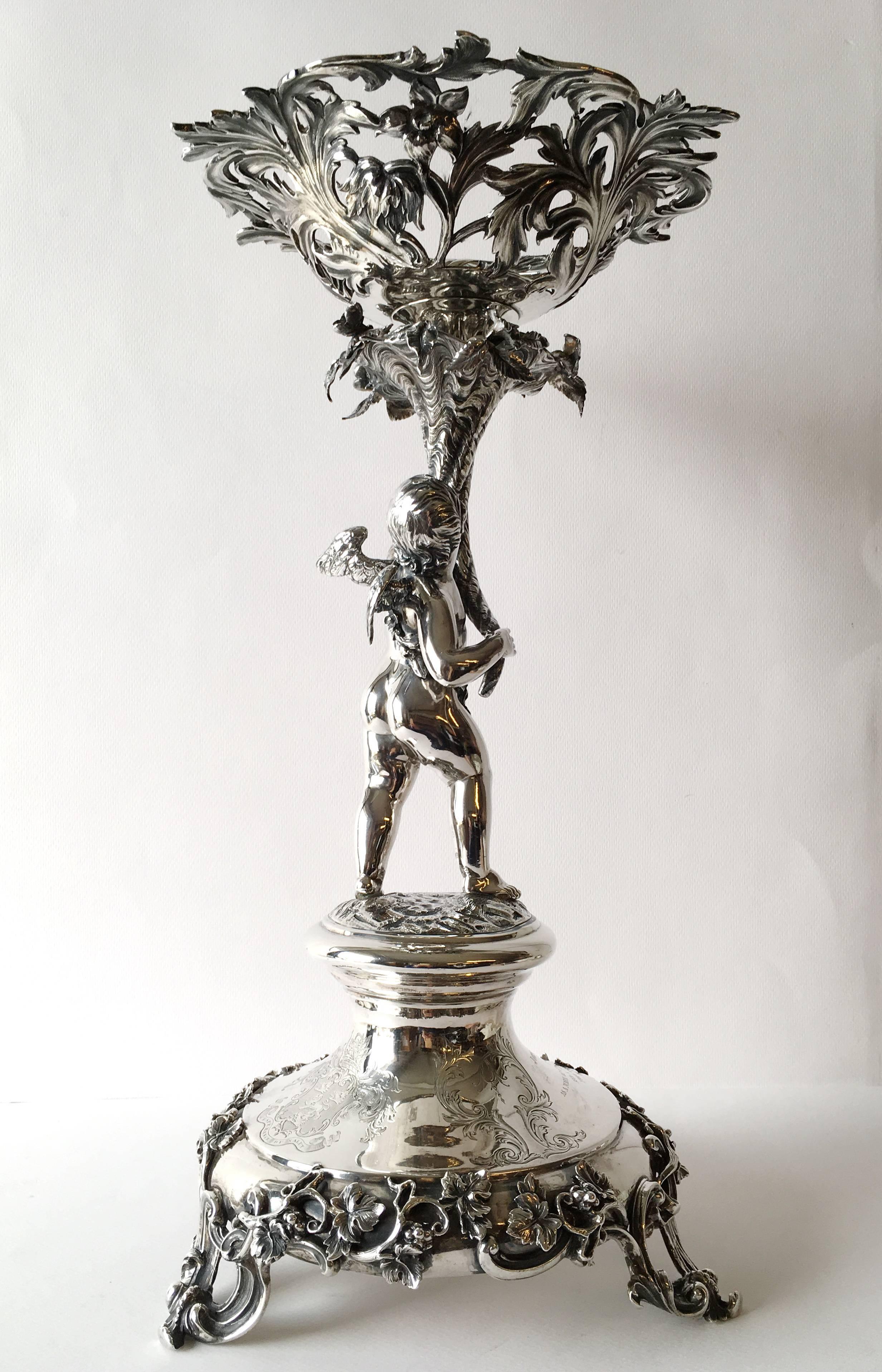 Raised on a high-domed and waisted base with cast grapevine, on three openwork scroll feet, surmounted by a winged putto having a draped garland of flowers around him holding a cornucopia edged with flowers, supporting an openwork detachable dish of
