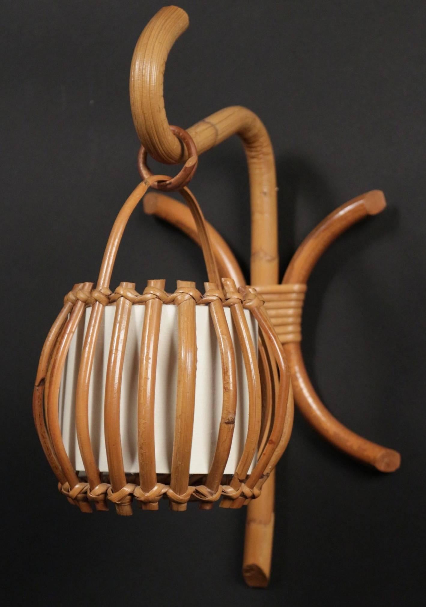 Pair of 1960s rattan "Lantern" sconces attributed to Louis Sognot.
Consist of a central rod underlined by two curved rattan sticks. On the top of the sconce a rattan buckle holds a cage shape lampshade dressed inside with cream white