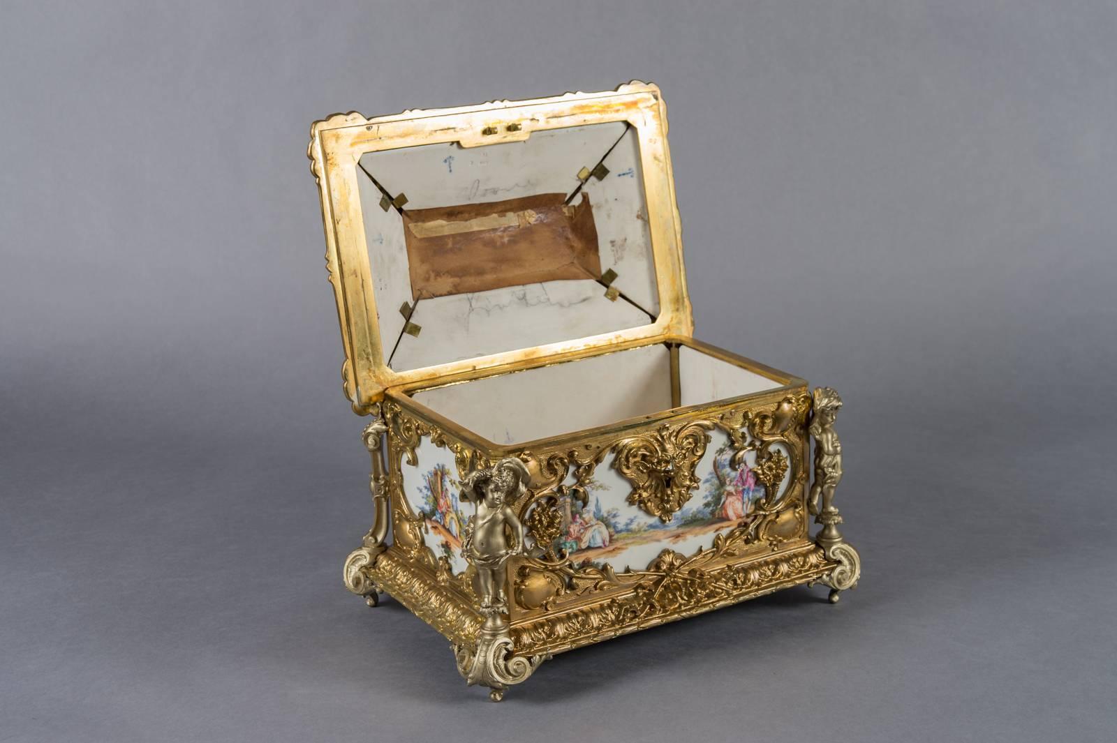 A large and impressive Berlin gilt and silvered bronze mounted porcelain casket. Surmounted by a rocaille handle flanked by putti herms, the cover applied with four silvered cornucopia, painted all over in the manner of Watteau with courting
