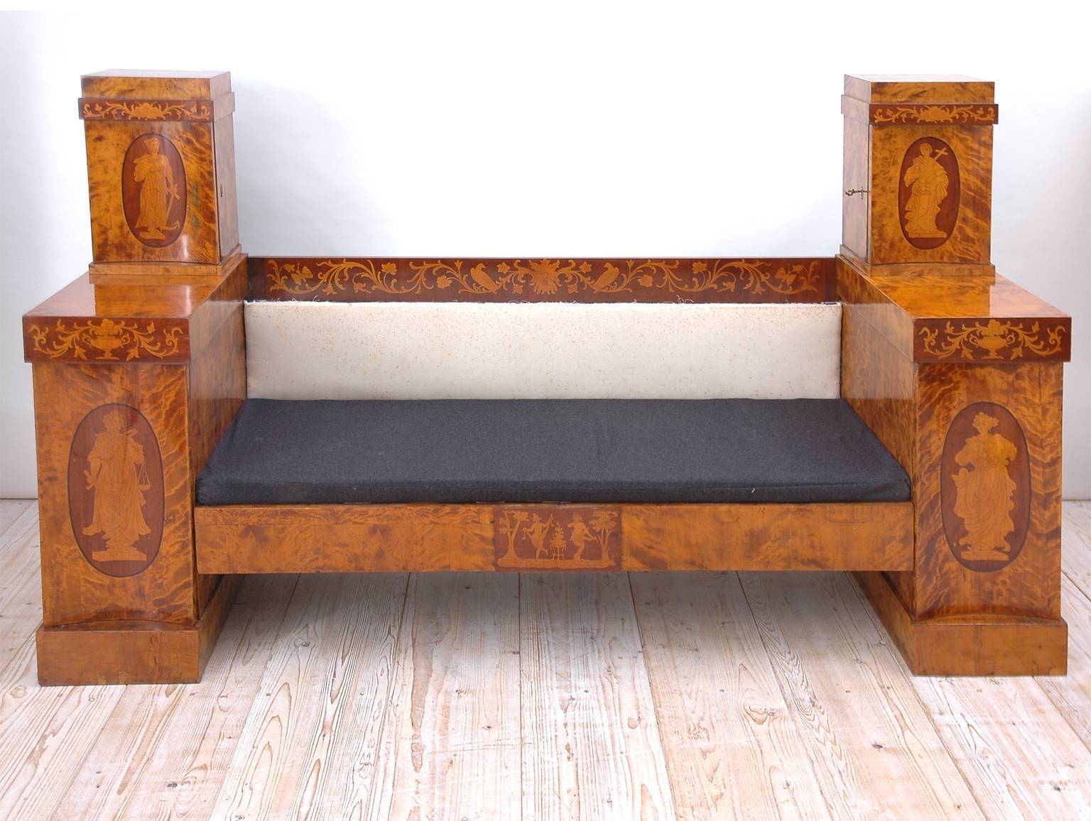 A Swedish Empire banquette/sofa in birch with marquetry embellishments which include foliage and urns and line-inlaid figures of the four cardinal virtues: Prudence, Justice, Fortitude and Temperance. Lower cabinets open to reveal drawers, while top