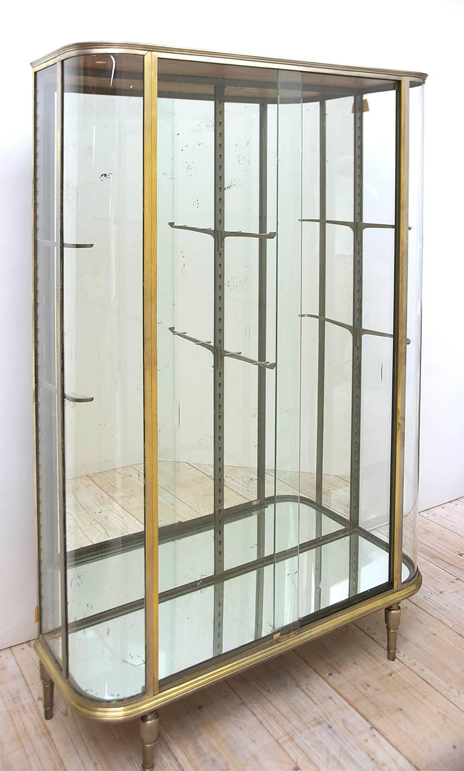 An exquisite Art Deco Vitrine with rounded glass and brass frame, circa 1920. Brass alloy conveys a silver quality more so then yellow gold. 
The most important elements of this vitrine are in tact which are the metal frame and the shaped glass on