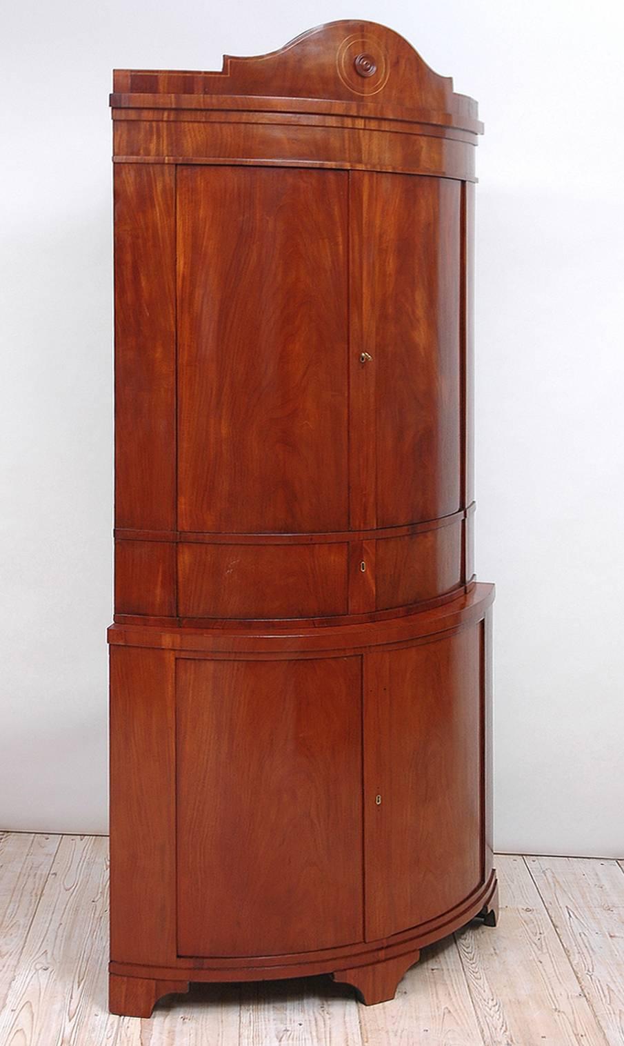 A large and very beautiful Karl Johan Empire corner cupboard in mahogany with tiara-shaped top banded with satinwood inlays and resting on bracket feet. Features a storage drawer midway between two sets of bowed-front doors that provide ample