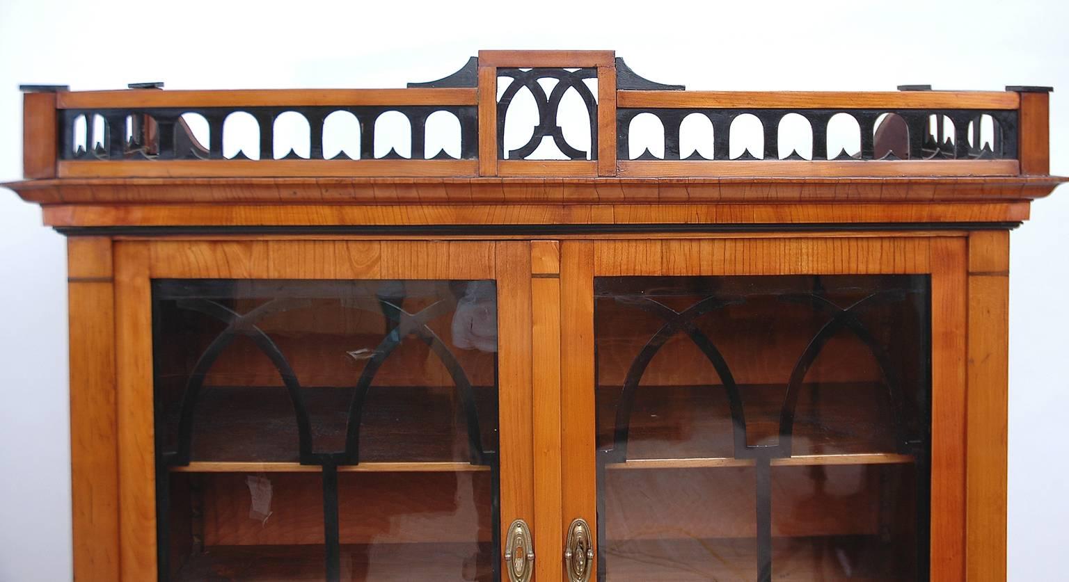 An Austrian Biedermeier vitrine in cherrywood with carved, open fretwork gallery, with ebonized details above crown molding. Arched lattice work on interior of glass doors is ebonized as are various moldings throughout. Offers display storage in