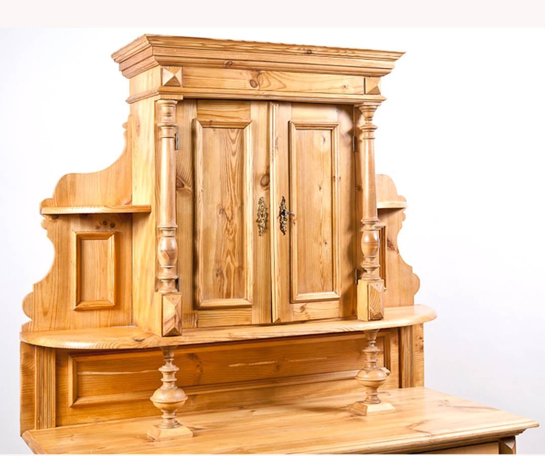 A Gründerzeit cupboard in pine with double-door upper cabinet flanked by open side shelves with paneled back-splash and resting on pillars, above base consisting of cabinet with two doors below two drawers. Full, turned columns give this charming