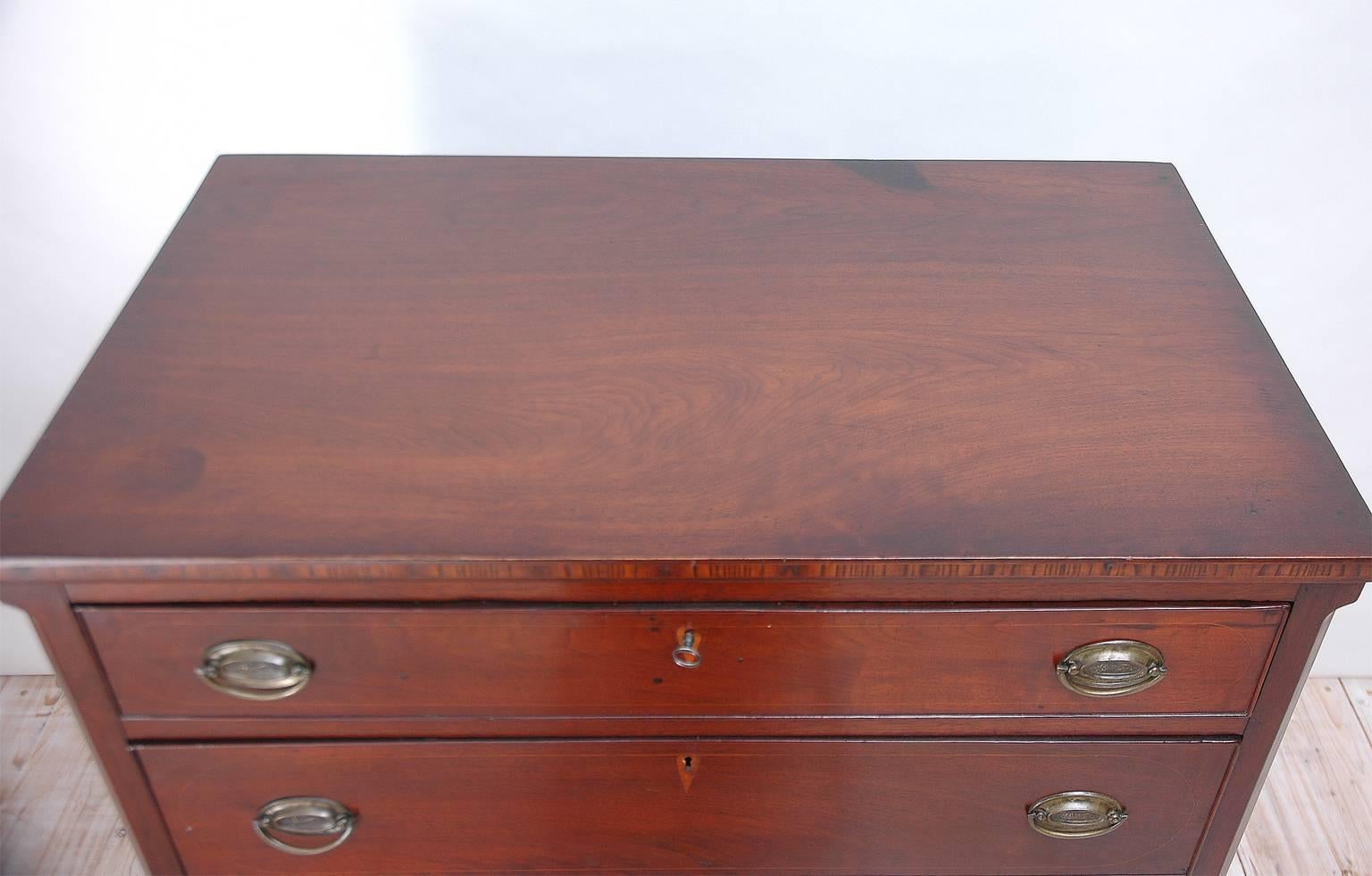A Hepplewhite chest in mahogany with four graduated drawers with inlaid key plates and original pulls depicting cornucopias. Decorative elements include line inlays around drawers, banding along top edge, and an inlaid pinwheel on apron, with cyma