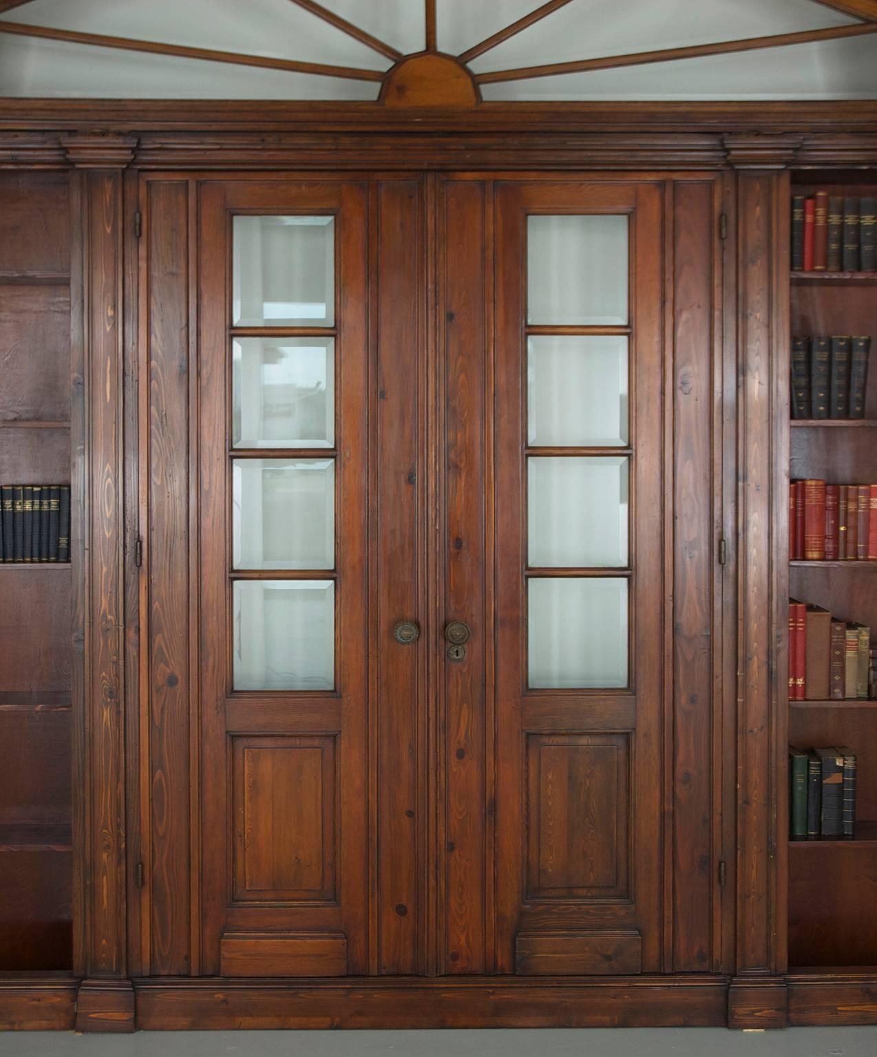 An impressive and large architectural pass-through or wall unit with bookcases flanking each side of two entry doors, over which spans a beveled glass transom in the shape of a sunburst that is encased in a rectangular panel decorated with gesso