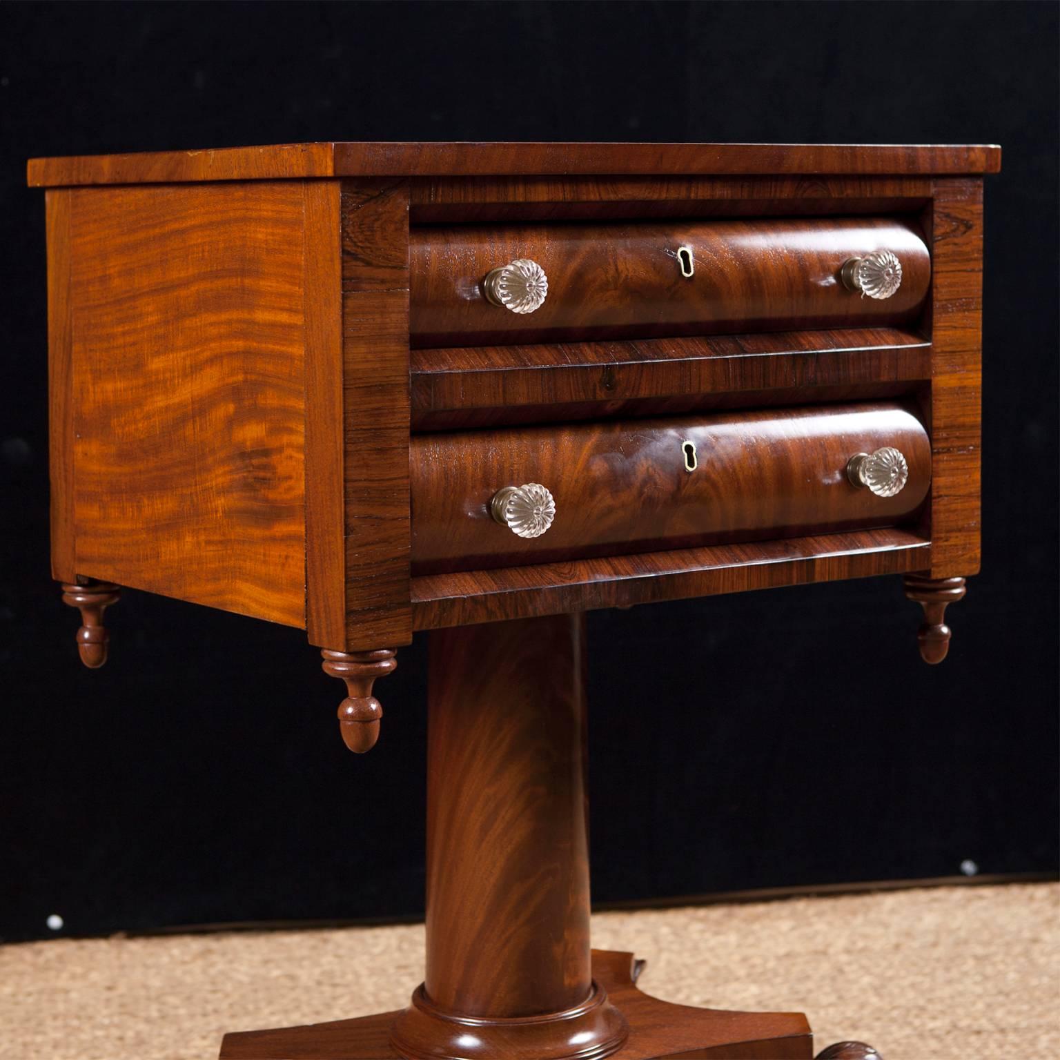 A neoclassical American Empire work table in mahogany and crotch mahogany with two drawers with convex fronts. Top rests on cylindrical column over a quatre-from base and terminating in carved lions' paw feet on casters, Philadelphia, circa