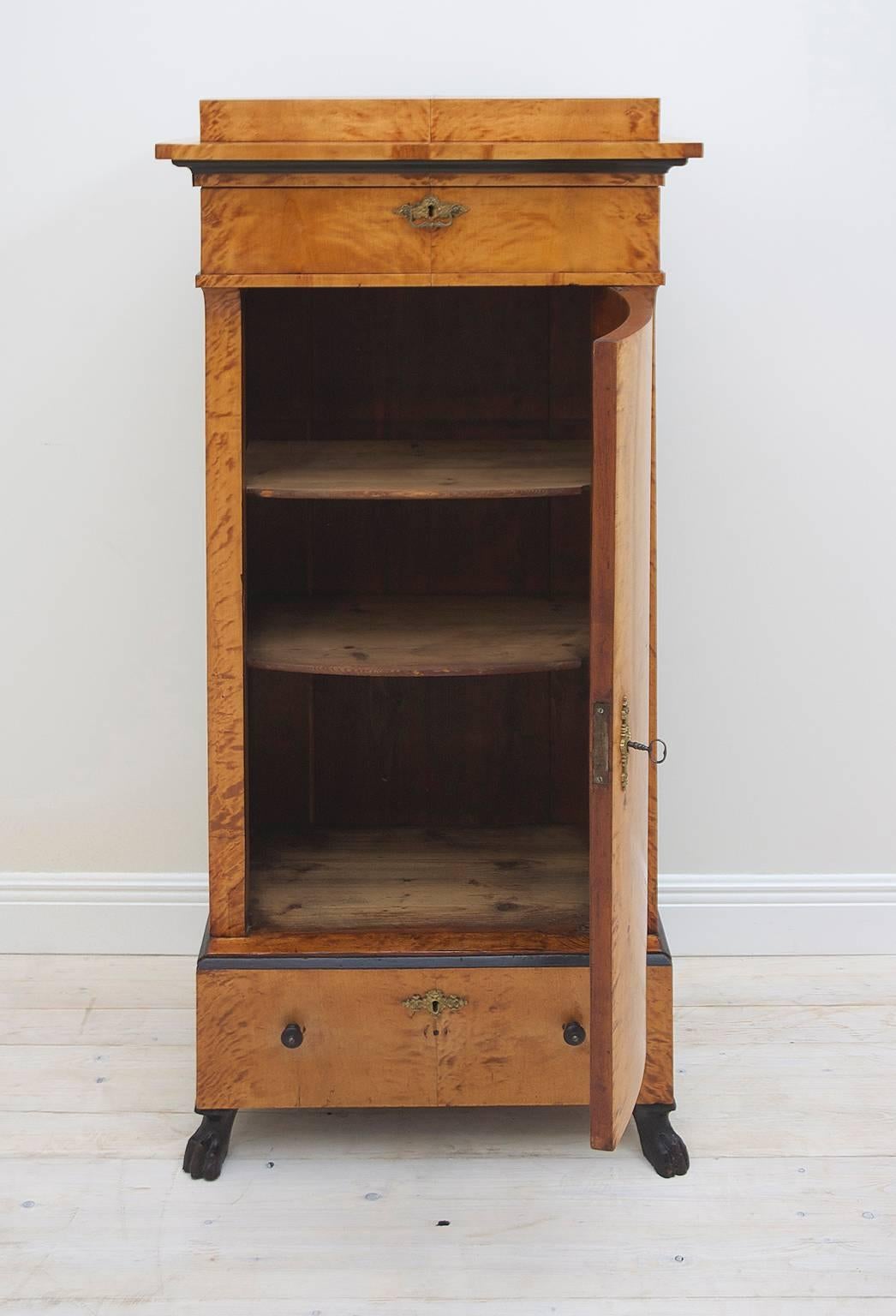 A Karl Johan Swedish Empire or early Biedermeier pedestal cabinet in birch offering two exterior drawers, one above and another below the central bowed door, with base resting on carved and ebonized lion's paw feet. Interior has three shelves,