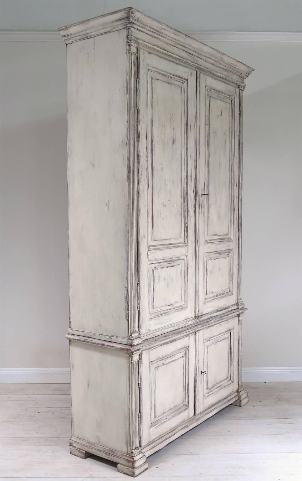 Tall Swedish four-door cupboard painted in the Gustavian manner. Cabinet features fluted pilasters framing a classical configuration of architectural raised door panels. Unit is to two parts. Bottom section features drawers while the top is